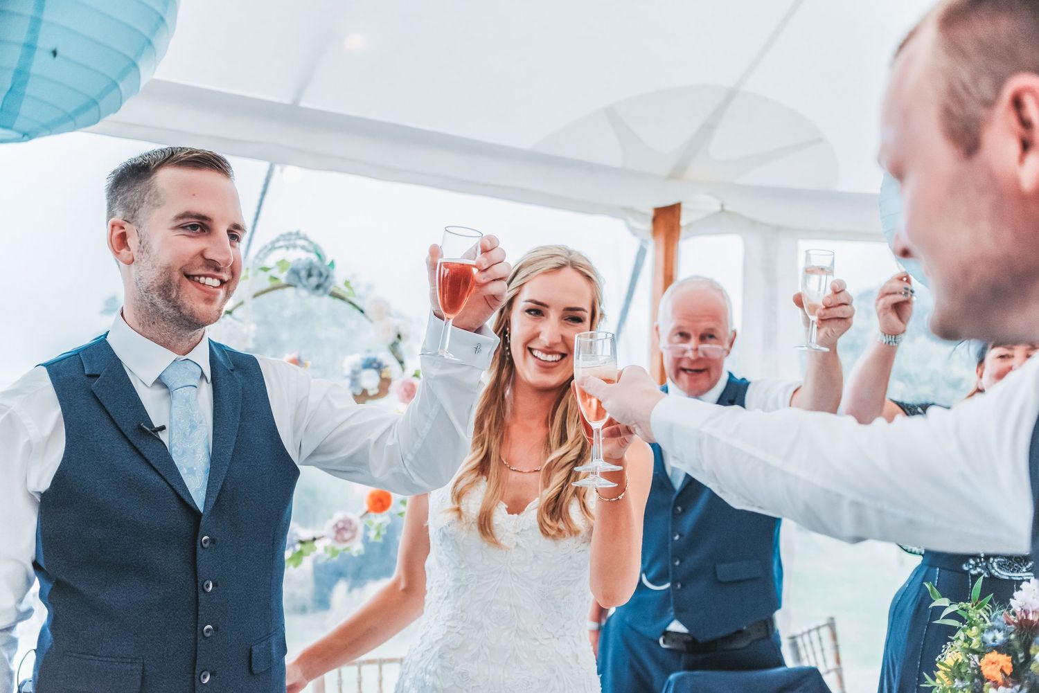 Wedding party celebrating with raised glasses under blue paper lanterns during a reception.