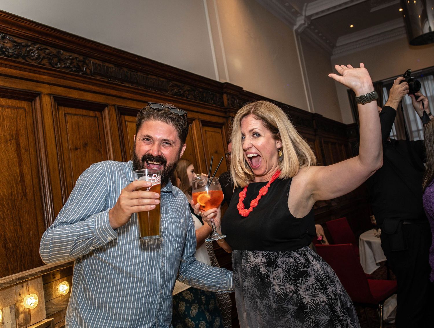 Party guests enjoying drinks and dancing at an indoor celebration with wood paneling.
