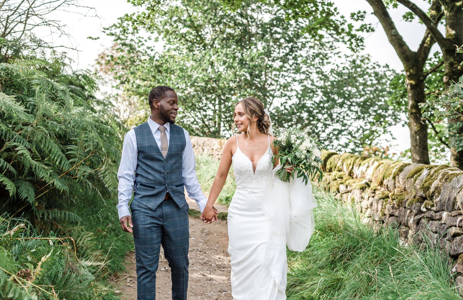 A couple walks hand in hand along a rustic path surrounded by greenery on their wedding day.