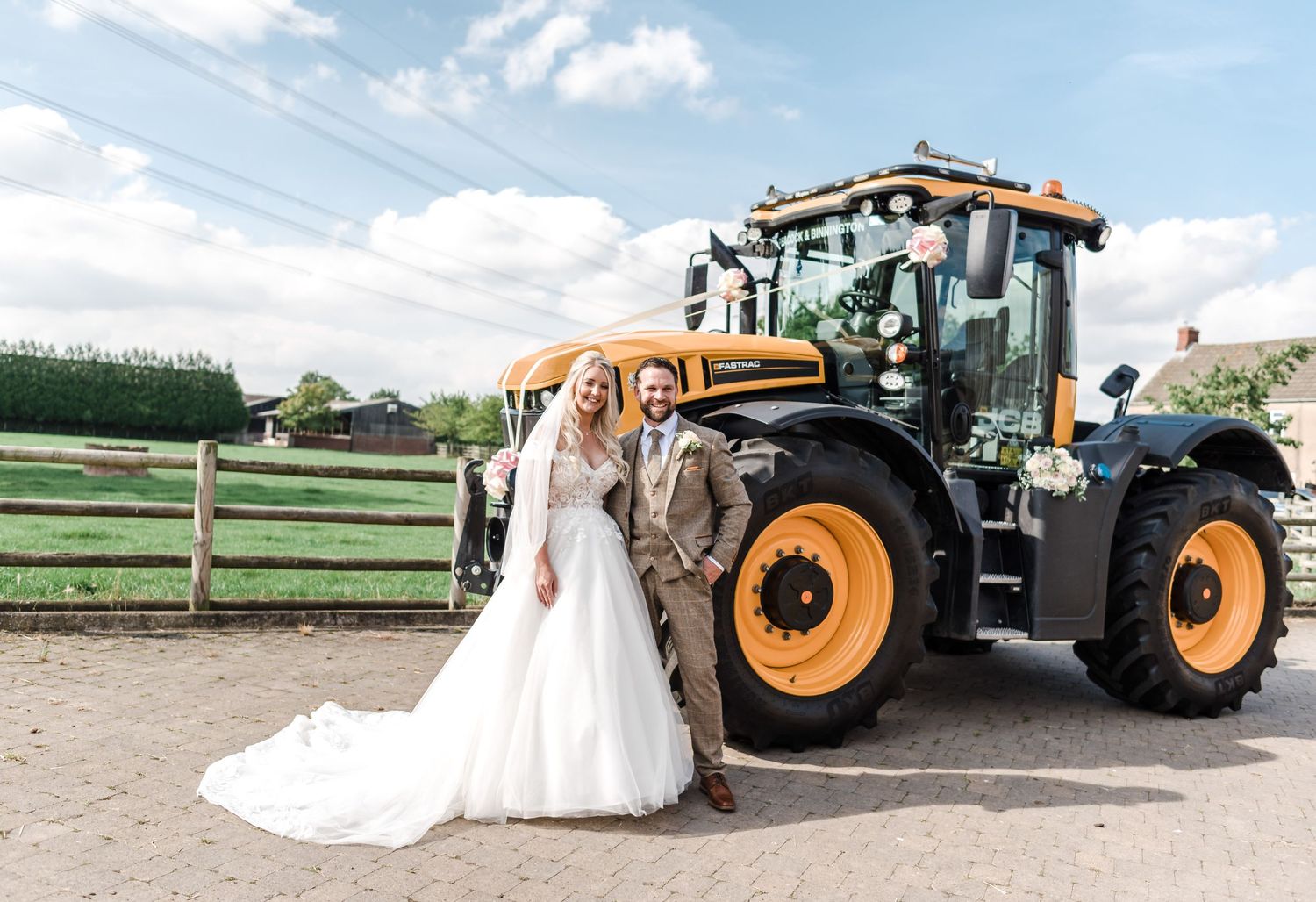 Wedding couple standing beside large yellow farm tractor in countryside setting.