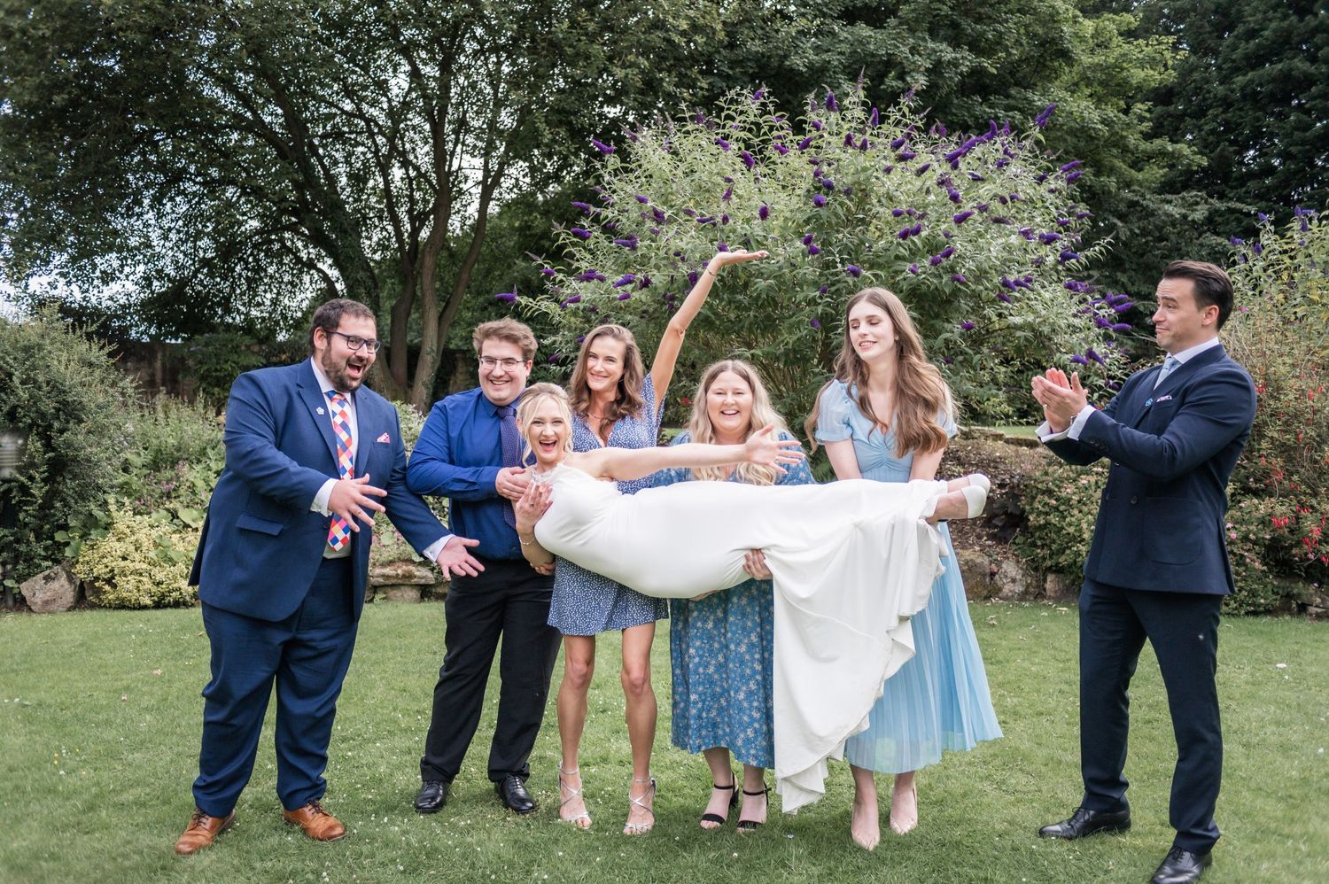 Wedding party playfully lifts and supports someone in a fun outdoor group photo.