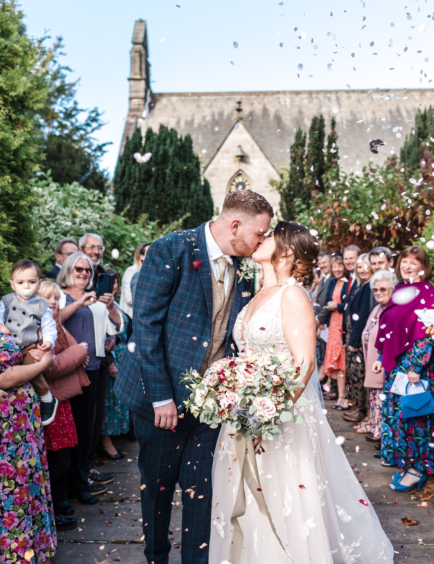 Wedding couple sharing kiss outside church while guests throw confetti.