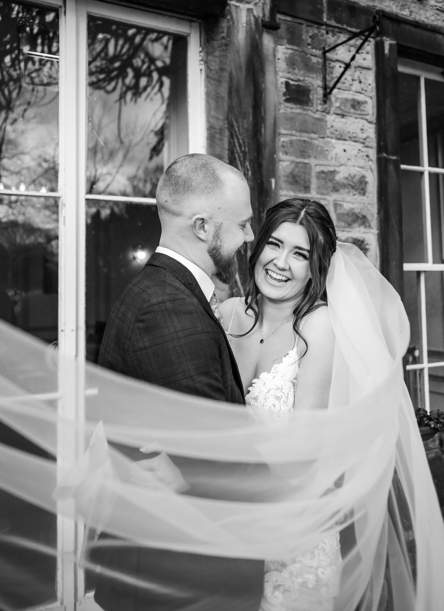 A romantic black and white wedding moment with a flowing veil against a brick backdrop.