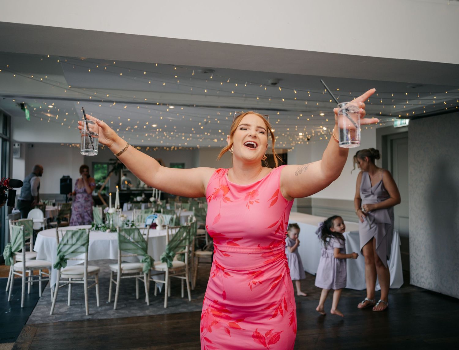 A guest in a bright pink dress celebrates joyfully with raised arms at an indoor event venue with twinkling lights.