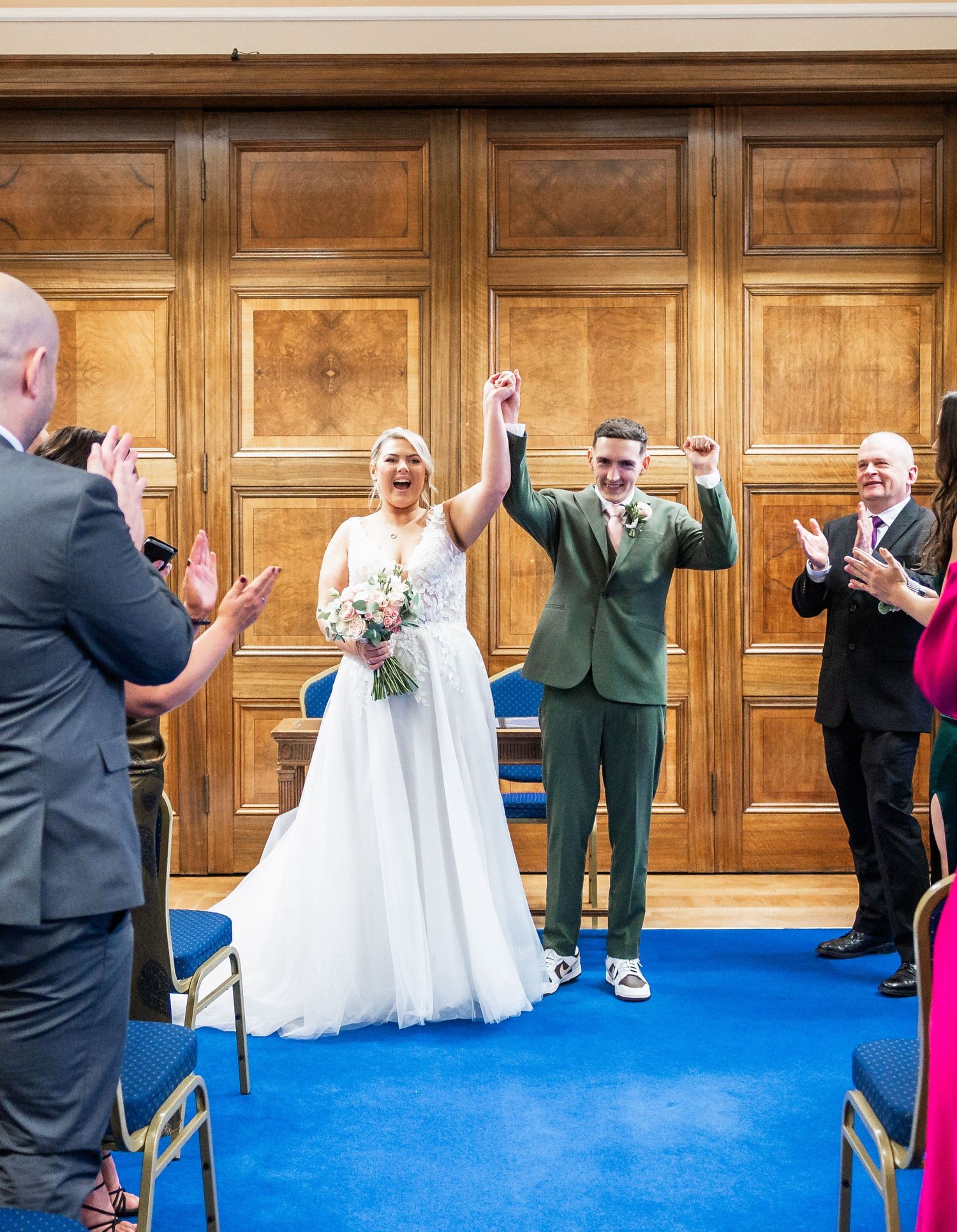 Newlyweds celebrate with raised arms as guests applaud in wood-paneled ceremony room.