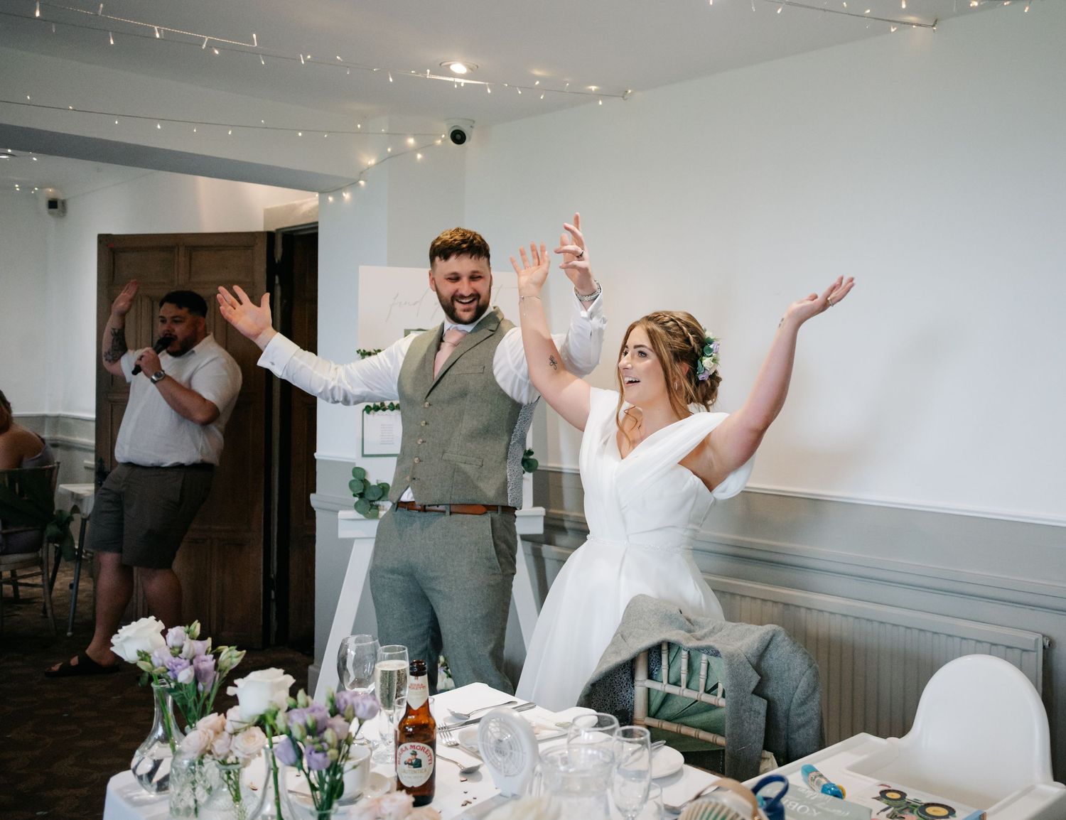 Wedding guests laugh and dance together with raised arms during a festive dinner reception celebration.