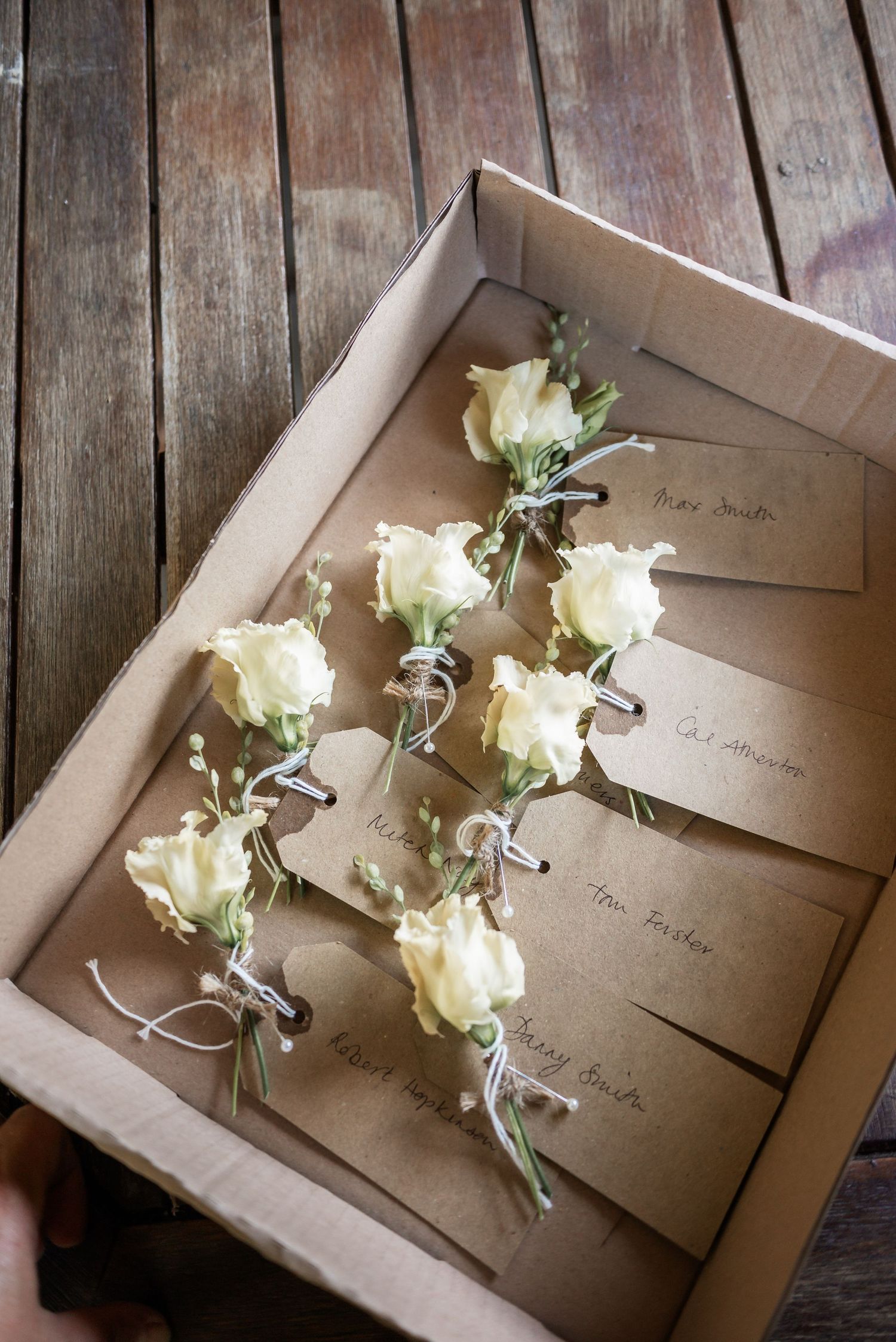 White rose boutonnieres with name cards arranged in a kraft paper box.