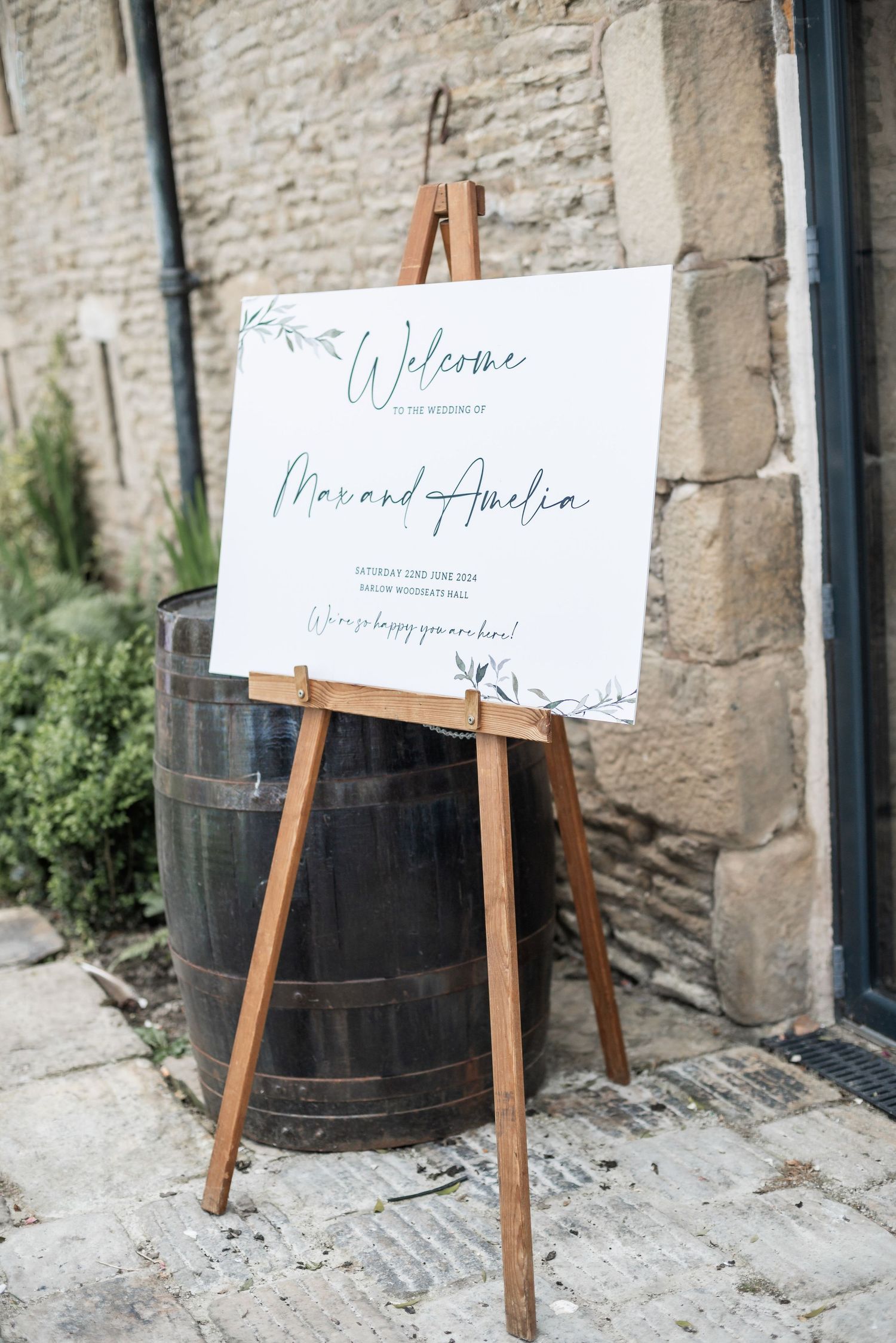 Wedding welcome sign on wooden easel next to vintage barrel against stone wall.