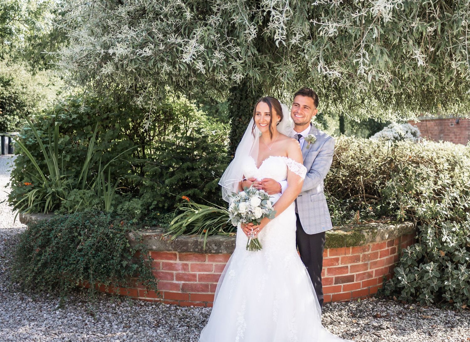 Couple poses with wedding bouquet in garden setting with brick wall.
