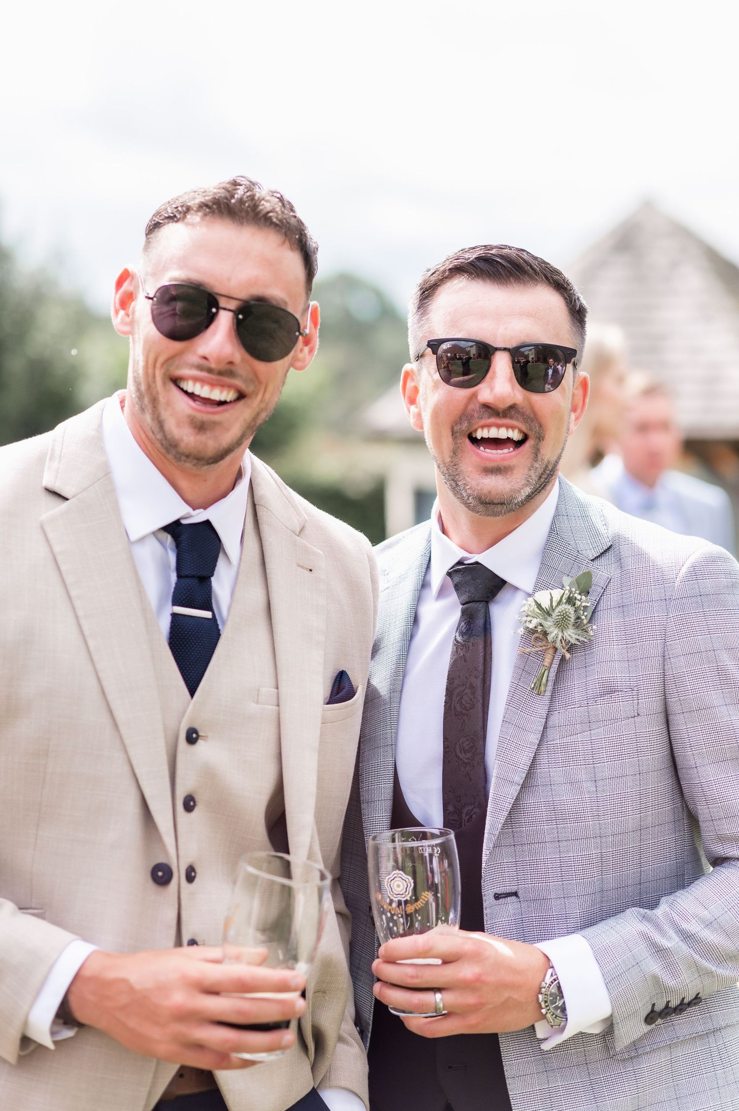 Two guests in sunglasses and suits share laughs while holding champagne glasses at an outdoor reception.