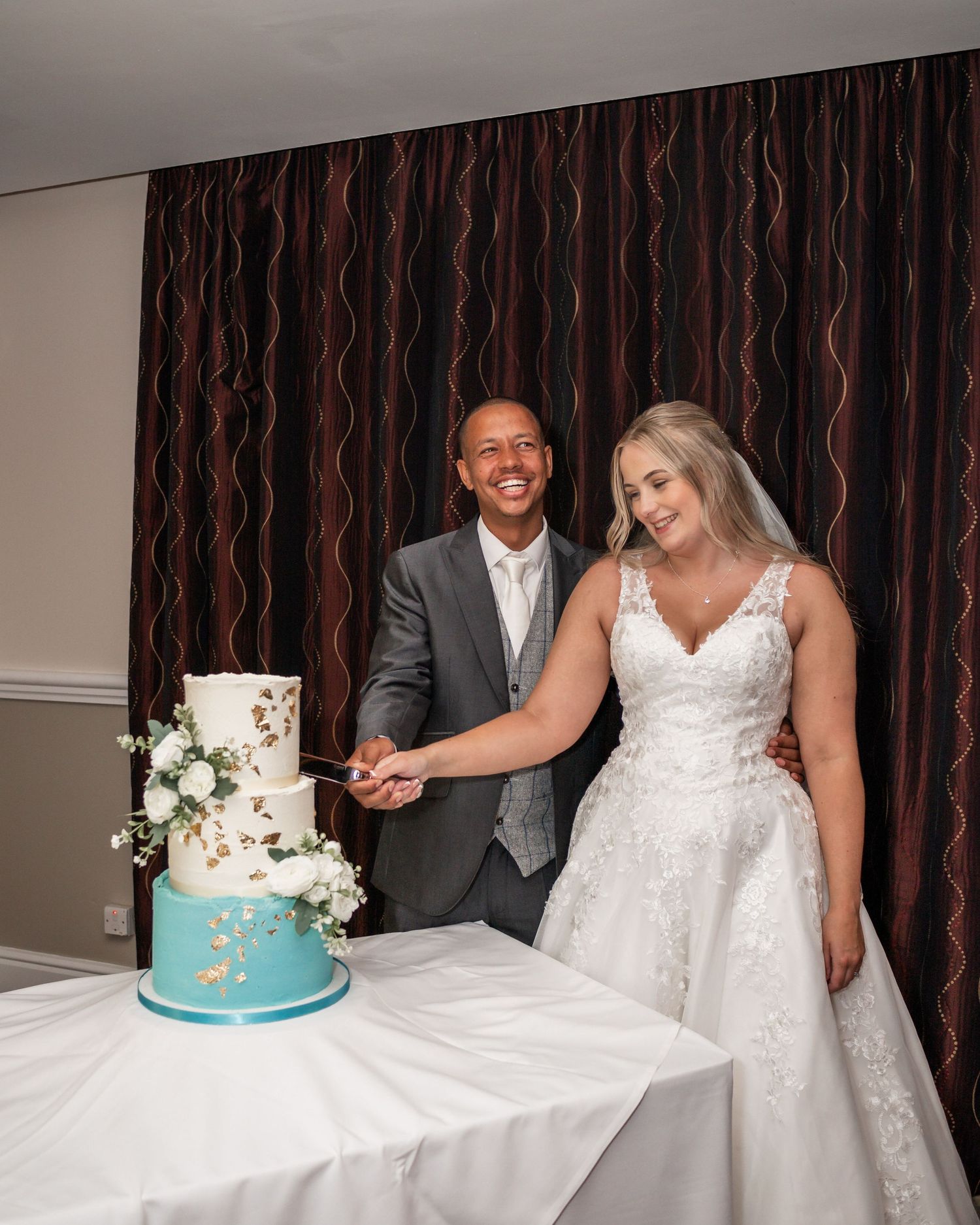 Newlyweds cutting their two-tier white and turquoise wedding cake against a striped curtain backdrop.