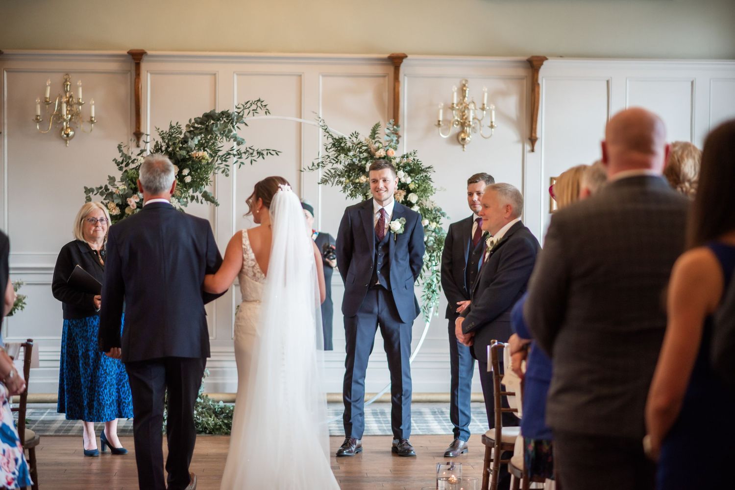Wedding ceremony in progress under elegant floral arch in classic indoor venue.