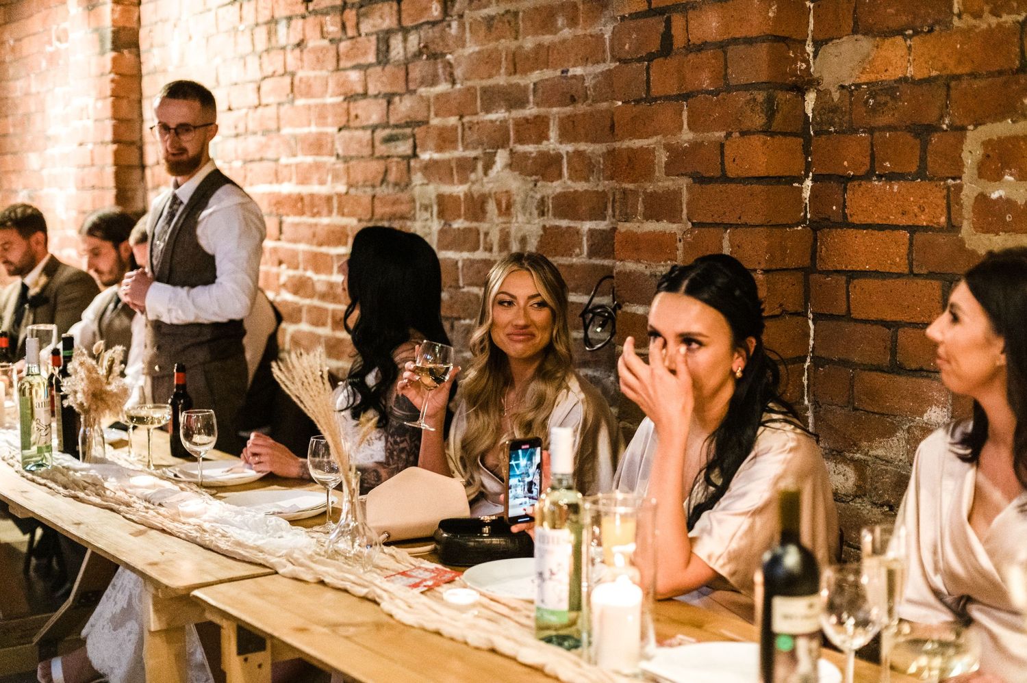 Wedding guests enjoying a reception dinner at a long table against exposed brick walls.