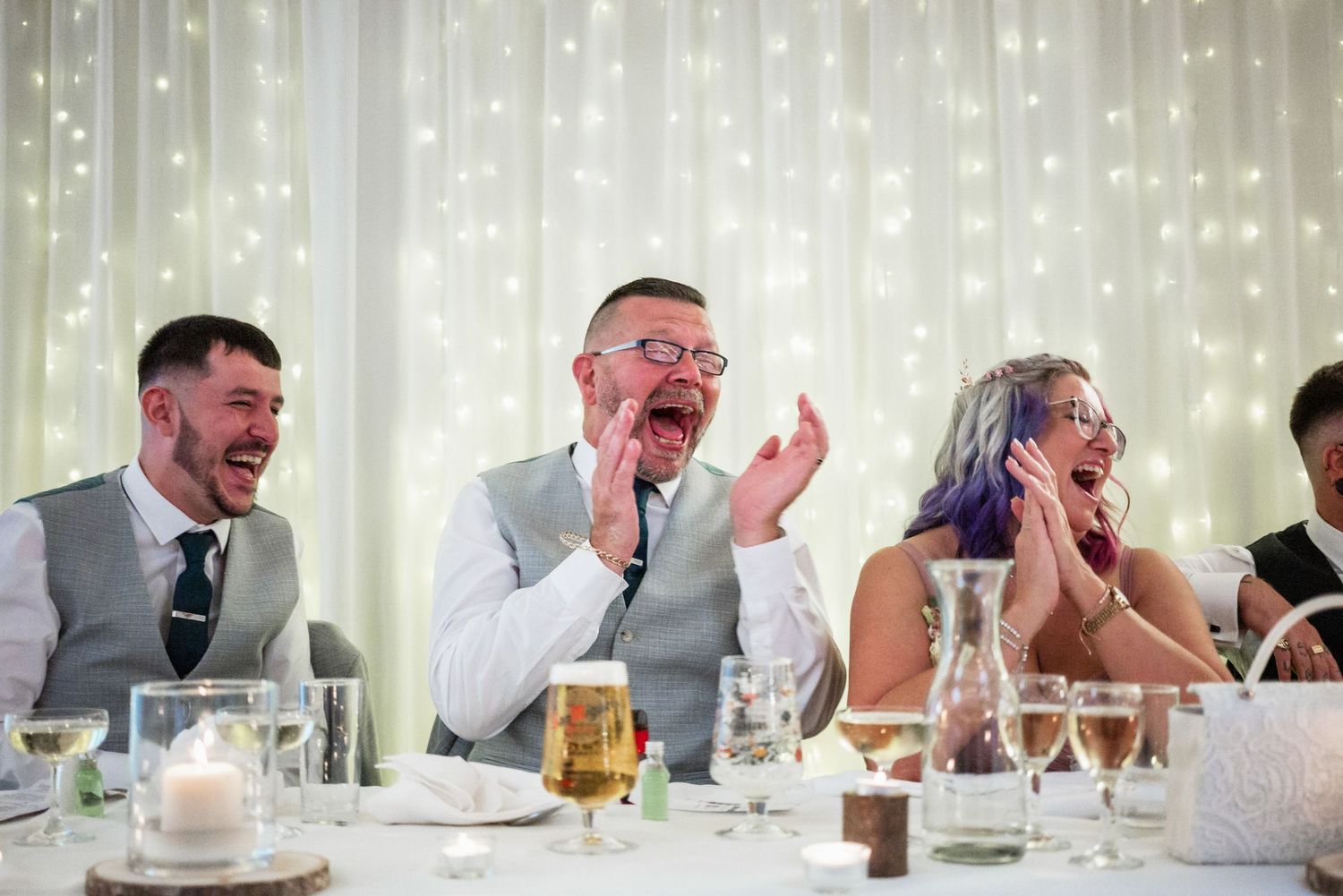 Wedding guests laugh joyfully at reception dinner with twinkle light backdrop.