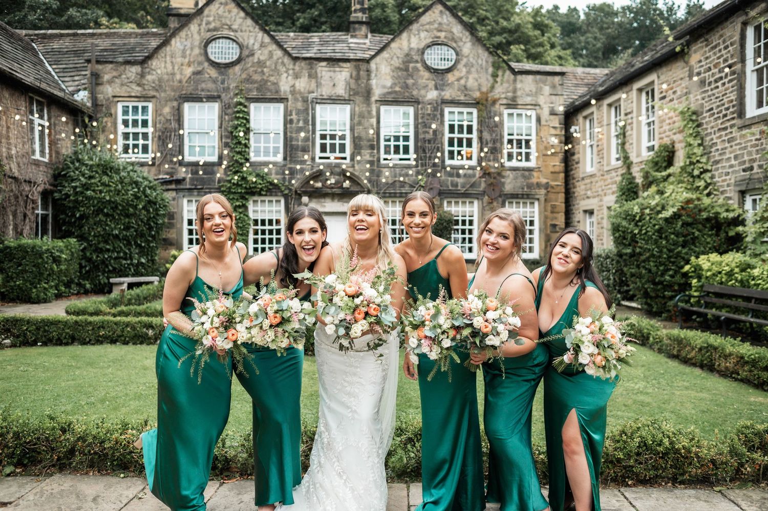 Wedding party in emerald green dresses pose with bouquets in front of historic stone building.