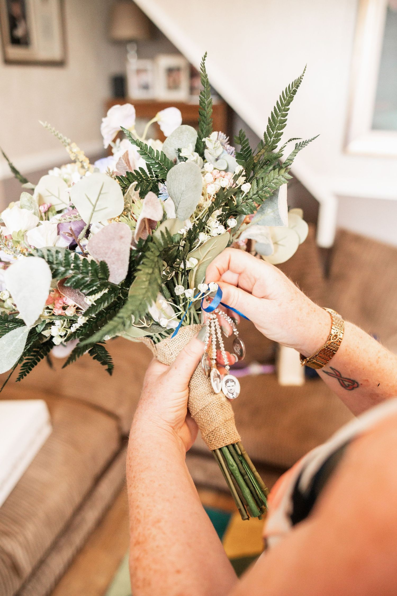 Hands adorned with jewelry arrange a delicate bouquet of white flowers and greenery against a cozy interior setting.