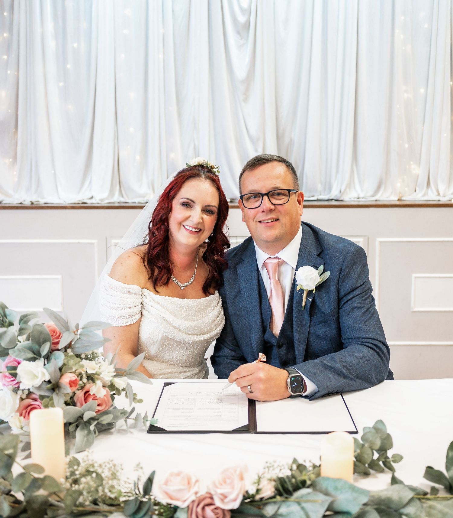 Newlyweds sign their marriage certificate at a decorated table with floral arrangements.
