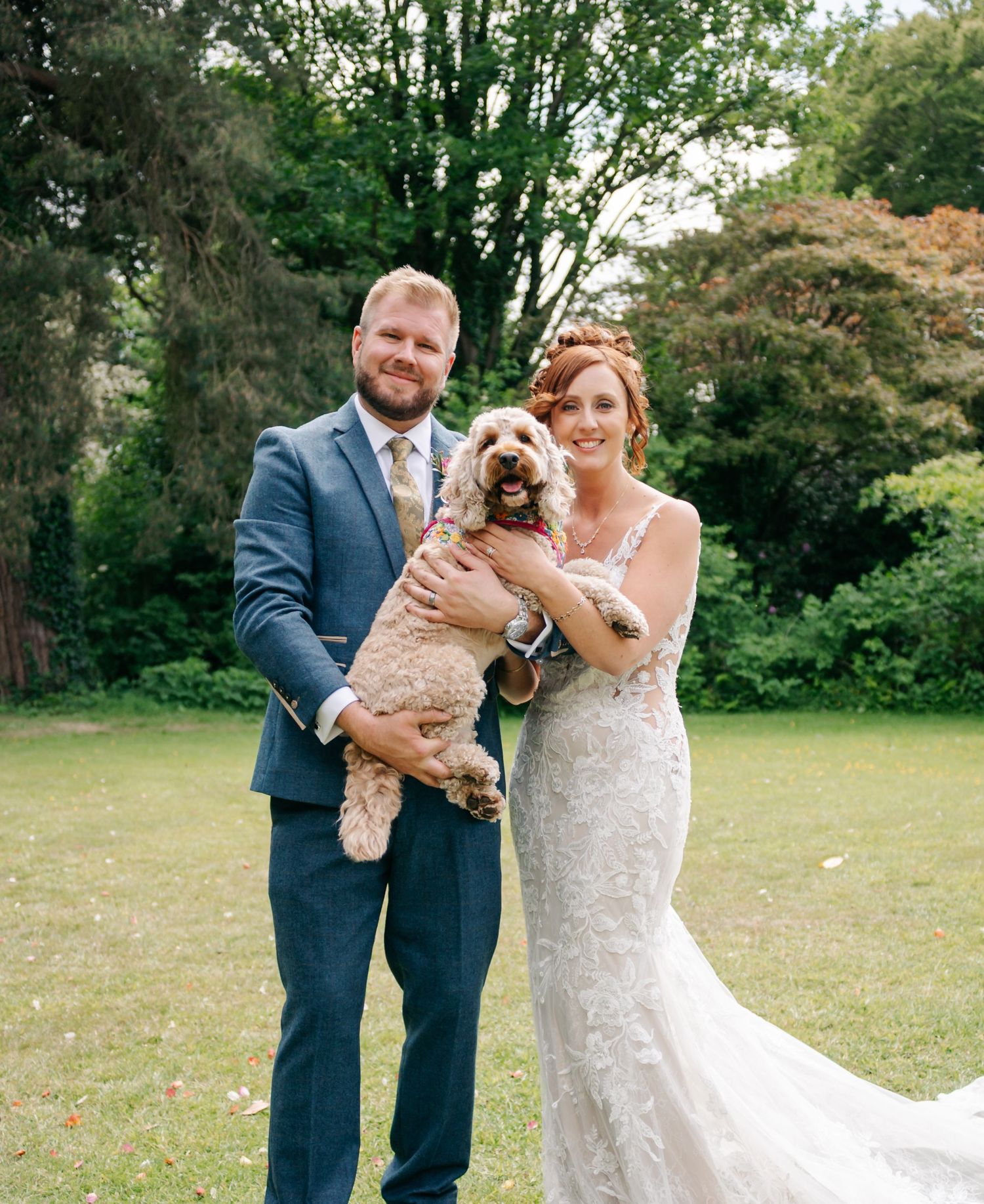 A couple in wedding attire holds their small dog in a sunny garden setting.