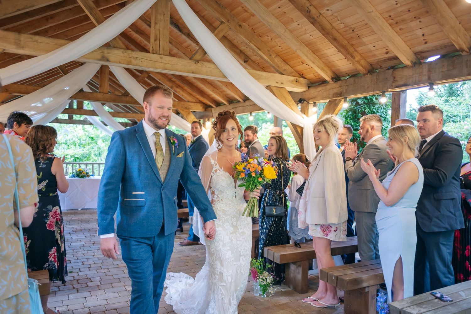 Guests watch as newlyweds celebrate during their rustic outdoor ceremony under wooden beams.