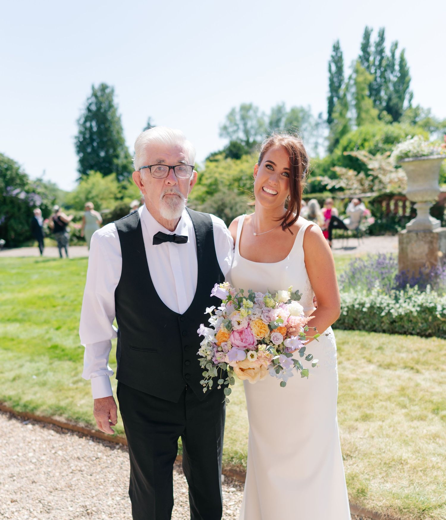 A couple poses together outdoors in wedding attire with floral bouquets on a sunny day.