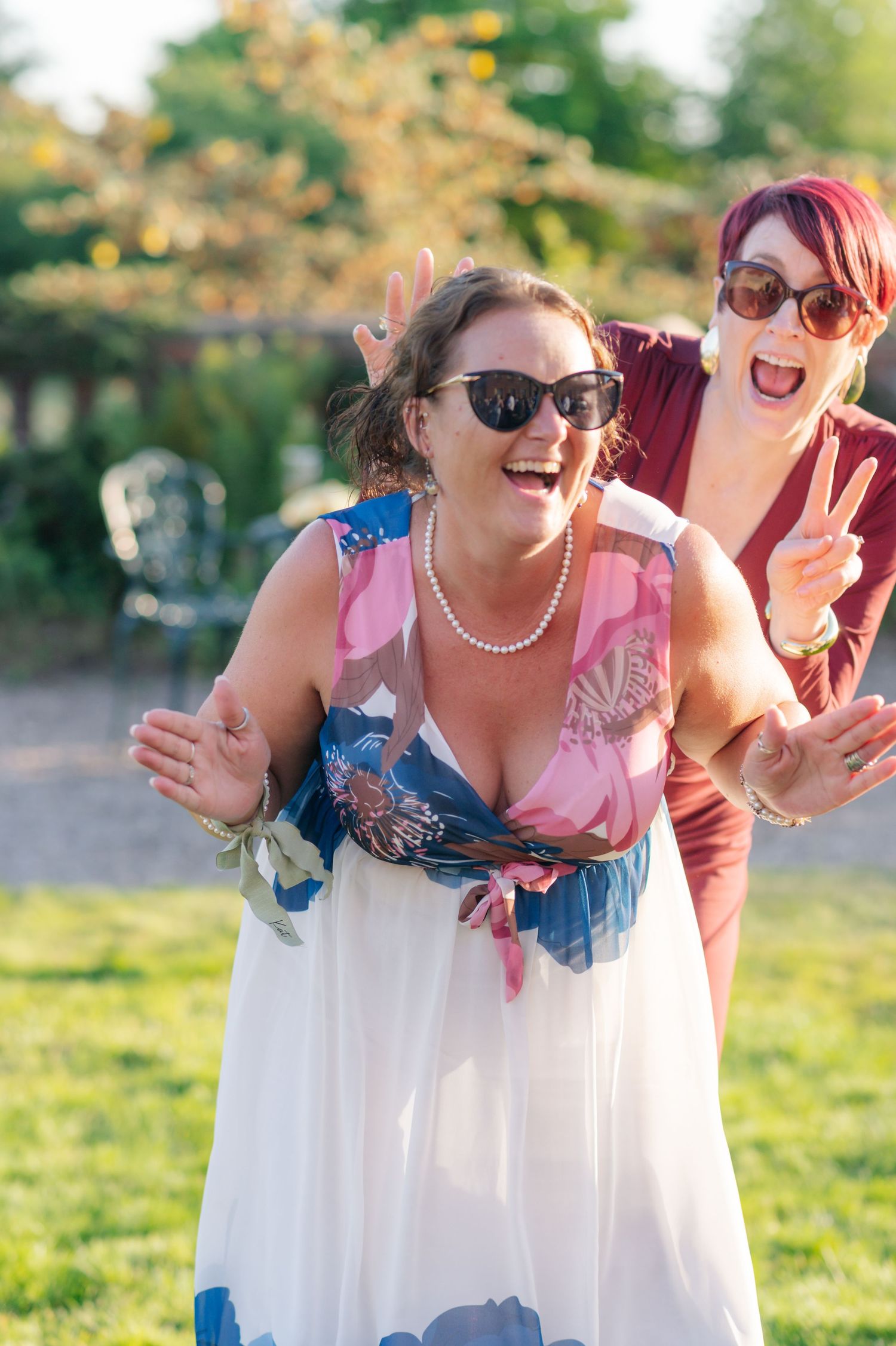 Friends laugh and pose together outdoors on a sunny day wearing colorful summer dresses and sunglasses.
