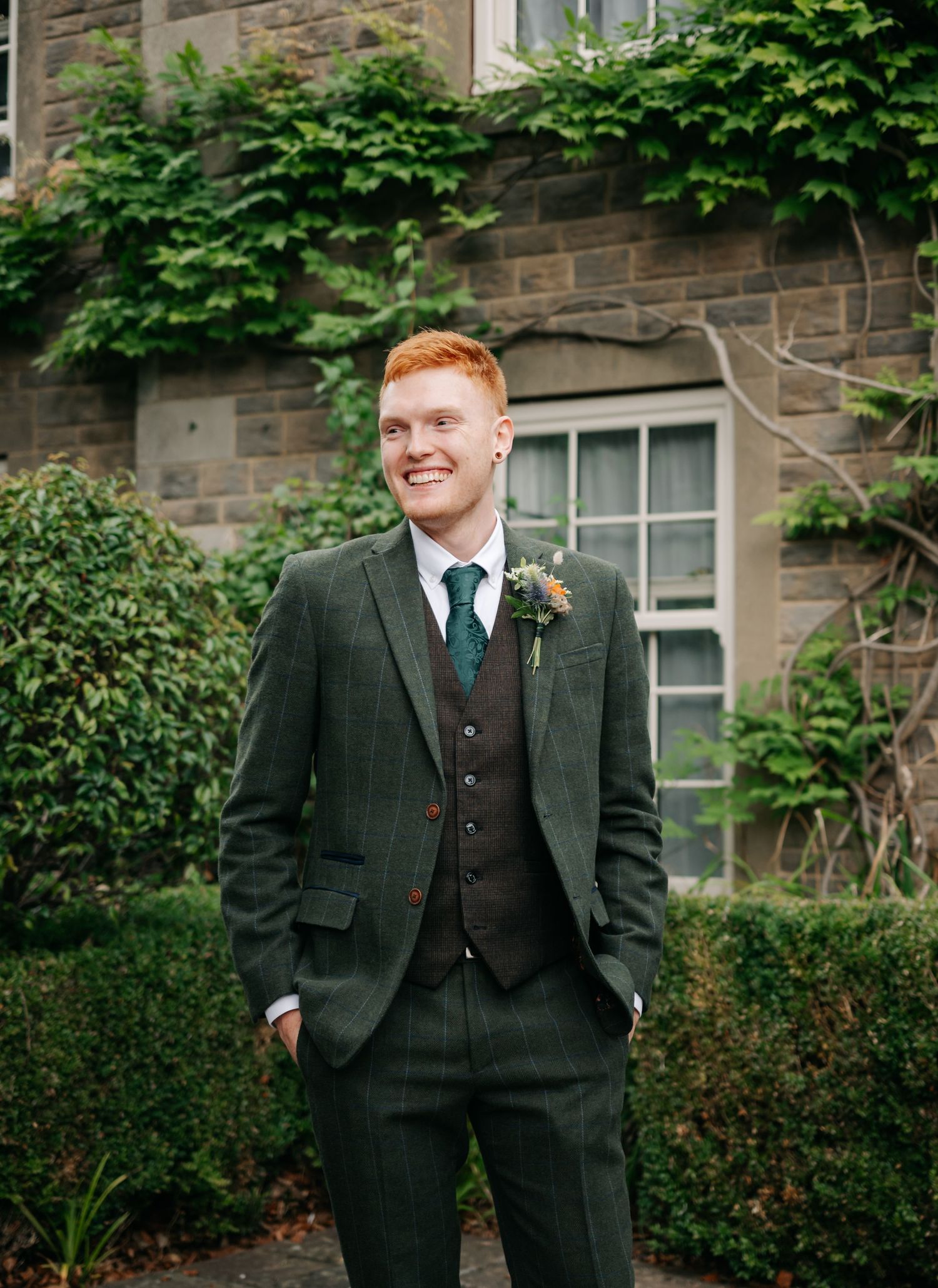 Groomsmen in dark green suits standing outside a stone building with ivy.