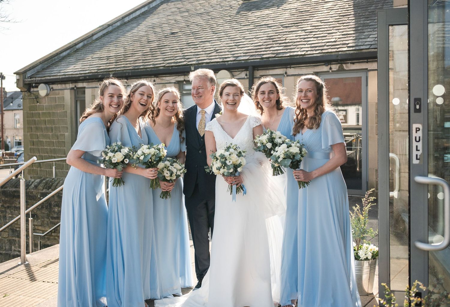 Wedding party in light blue bridesmaid dresses poses with bride in white gown outside brick building.
