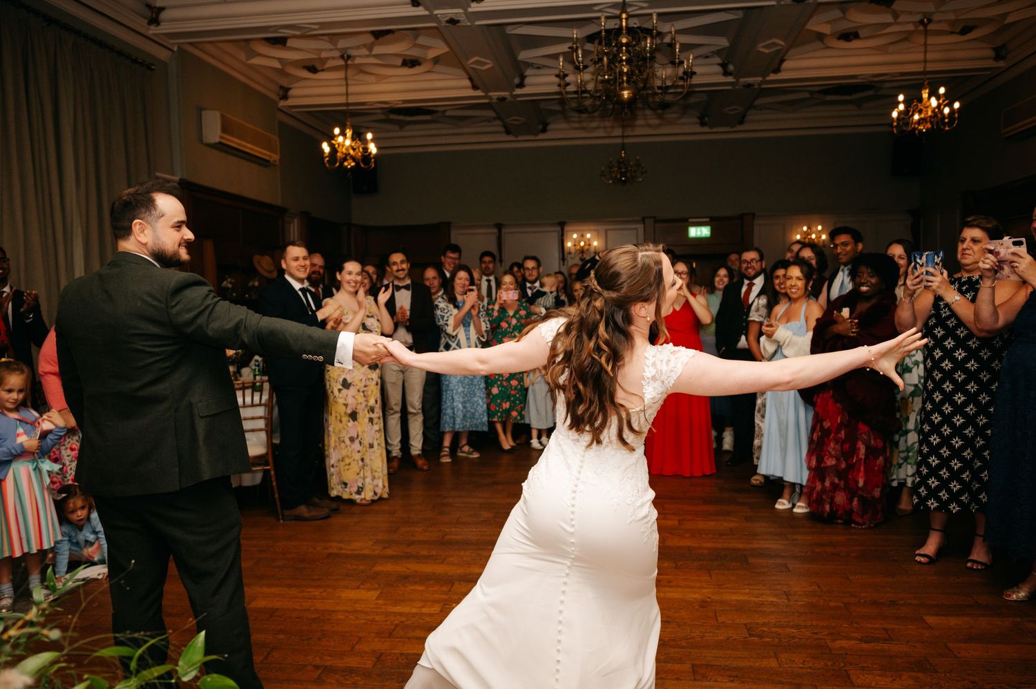 Bride and groom hold hands and dance in the middle of a wood-floored ballroom as wedding guests watch and cheer around them.