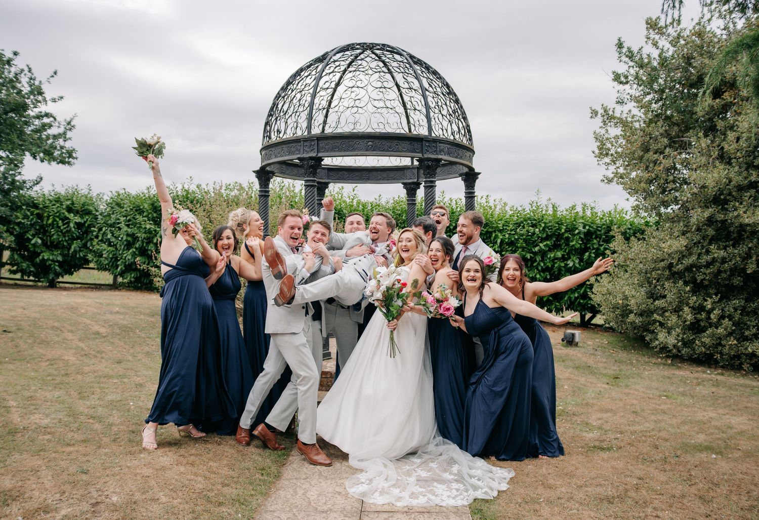 Wedding party poses under decorative metal gazebo in outdoor garden setting.