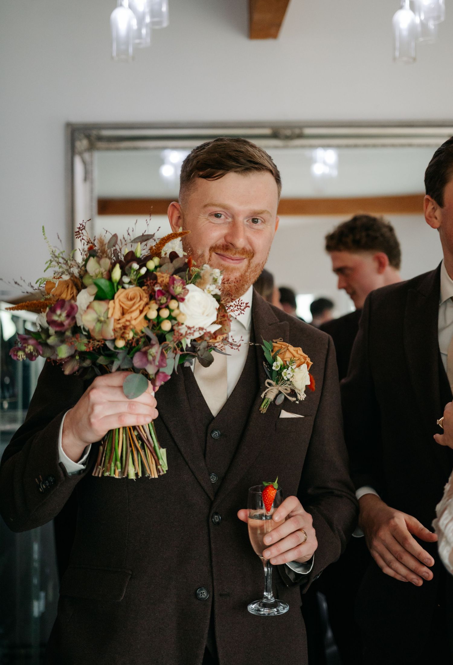 Wedding attendant in dark suit holds a peach and white floral bouquet while raising a champagne glass.