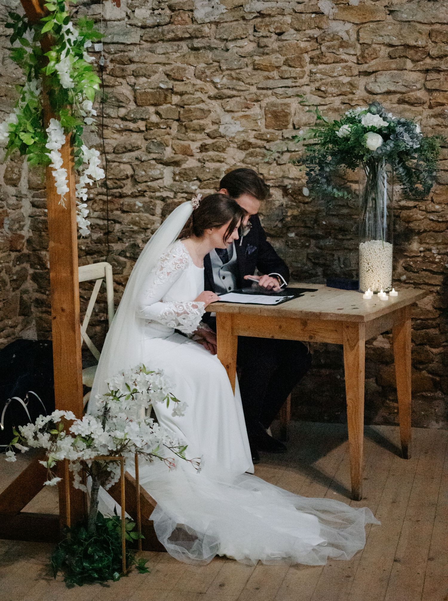 Wedding ceremony setup with rustic stone wall backdrop and wooden benches decorated with floral arrangements.