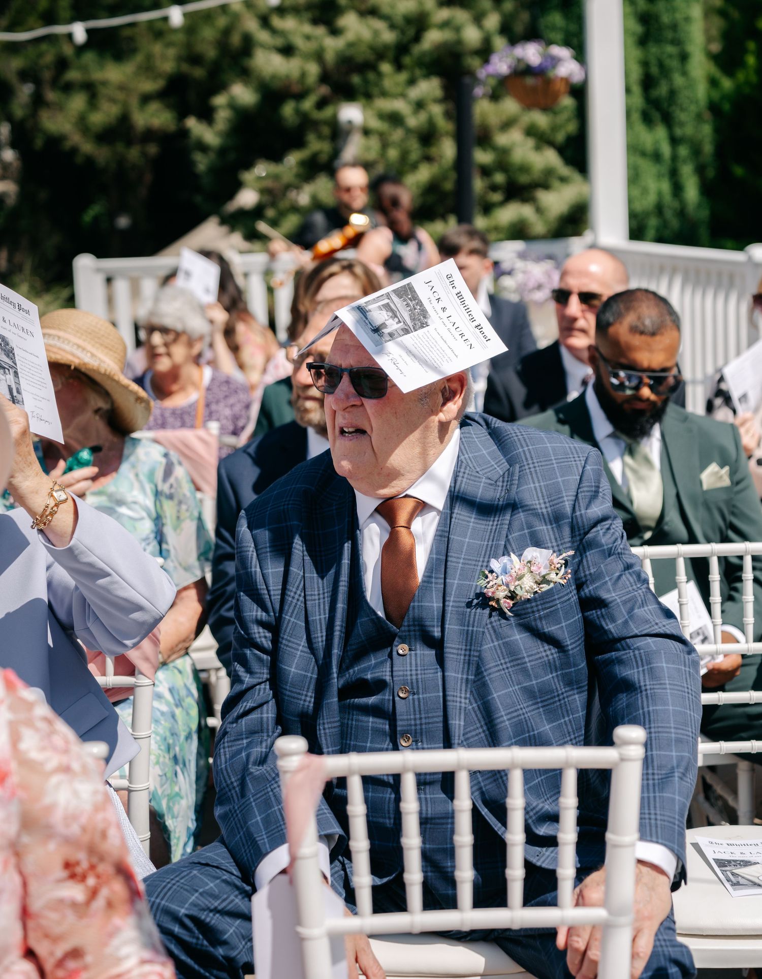 Dapper wedding guest in blue check suit using order of service as sun shade during outdoor ceremony.