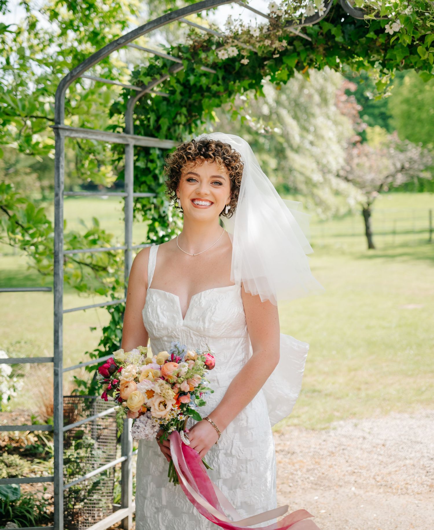 Outdoor garden wedding scene with white dress, floral bouquet and flowing veil under natural archway.