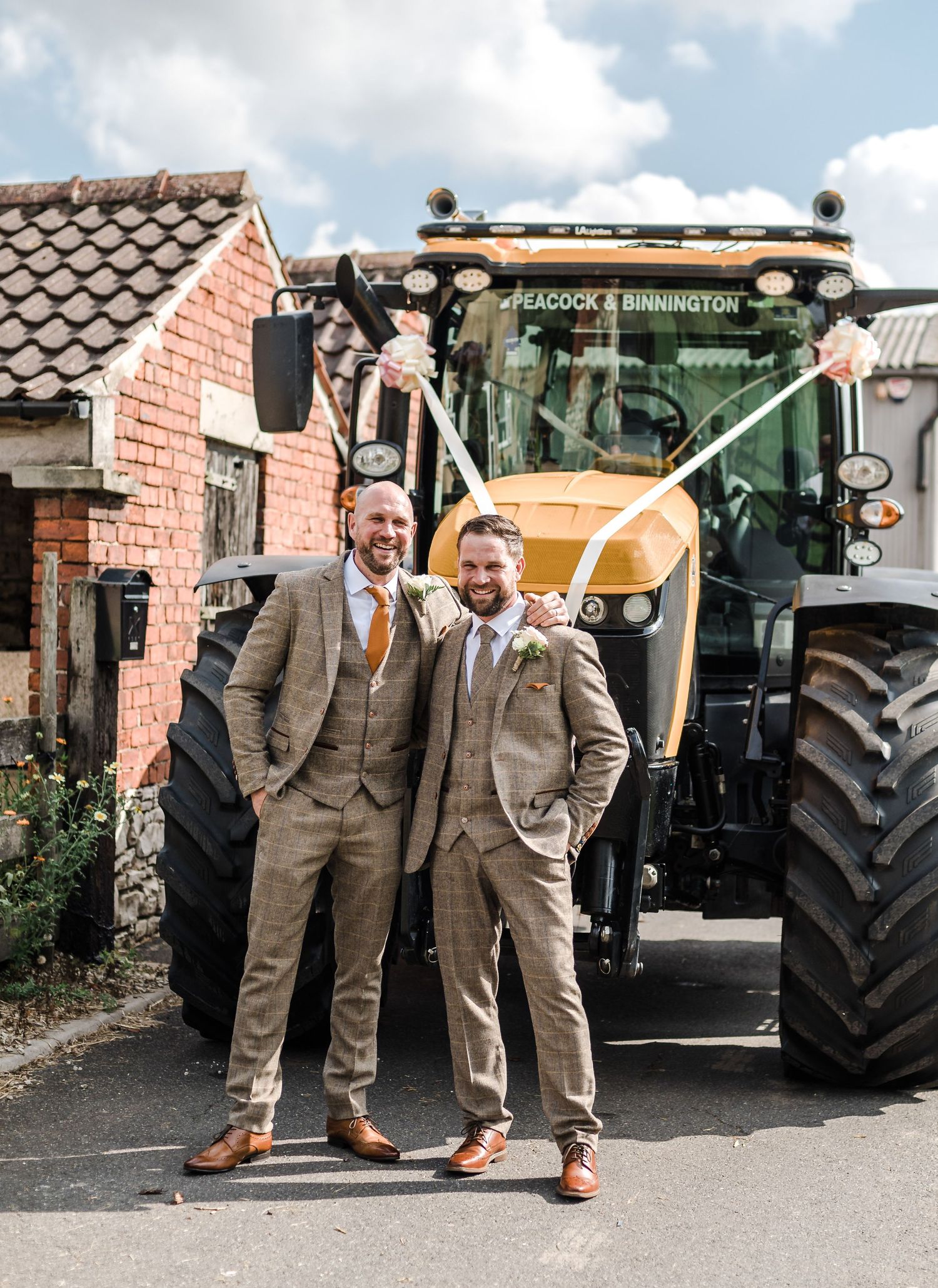 Two people in tweed suits pose in front of a large agricultural tractor.