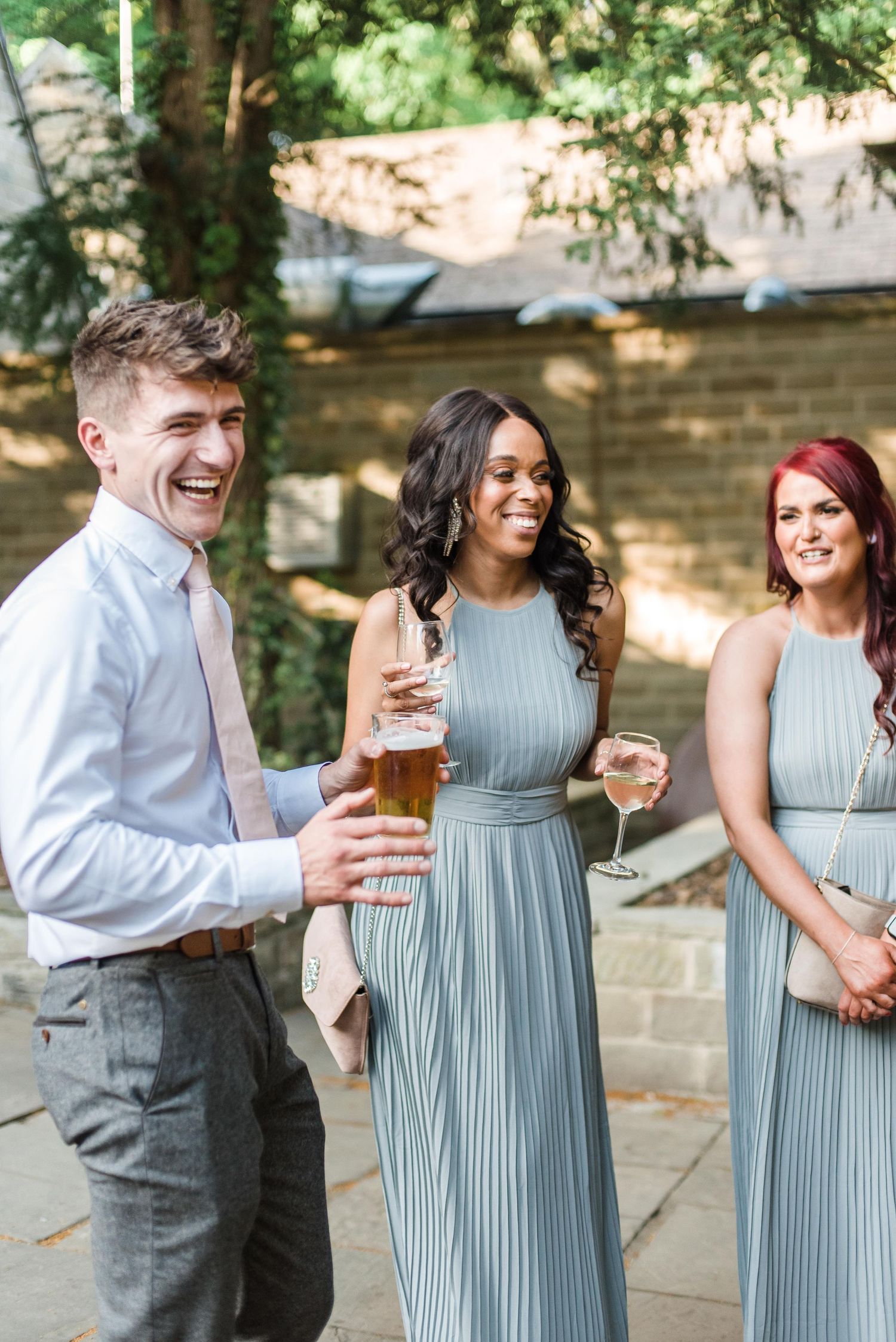 Wedding guests in grey and blue attire share drinks and laughter at an outdoor reception.