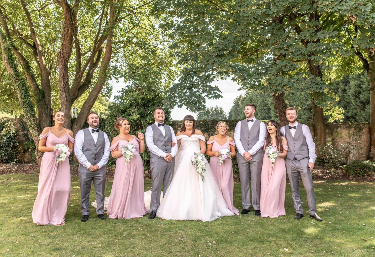 Wedding party in pink bridesmaid dresses and gray suits pose outdoors under trees for formal group photo.
