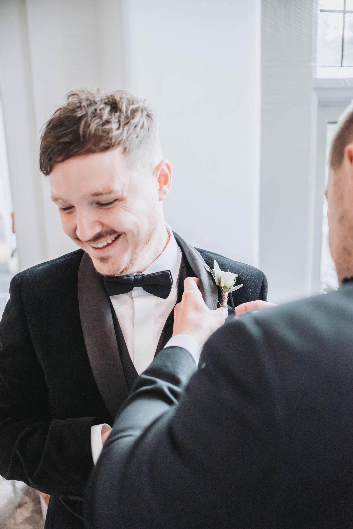 Groomsman helps adjust boutonniere on groom's black tuxedo lapel.