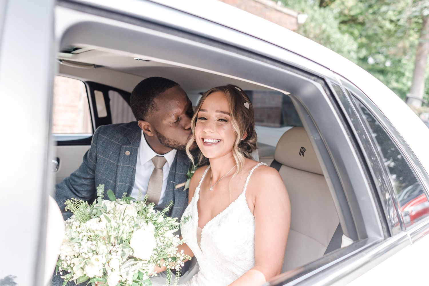 A happy couple shares a moment in their wedding car with a white floral bouquet.