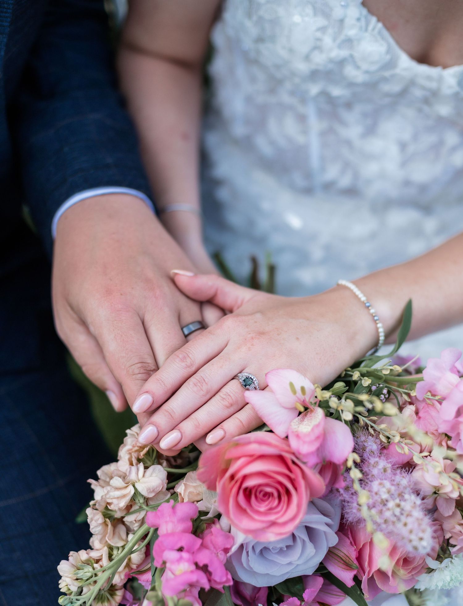 Close-up of wedding rings and hands on pink and purple floral bouquet.