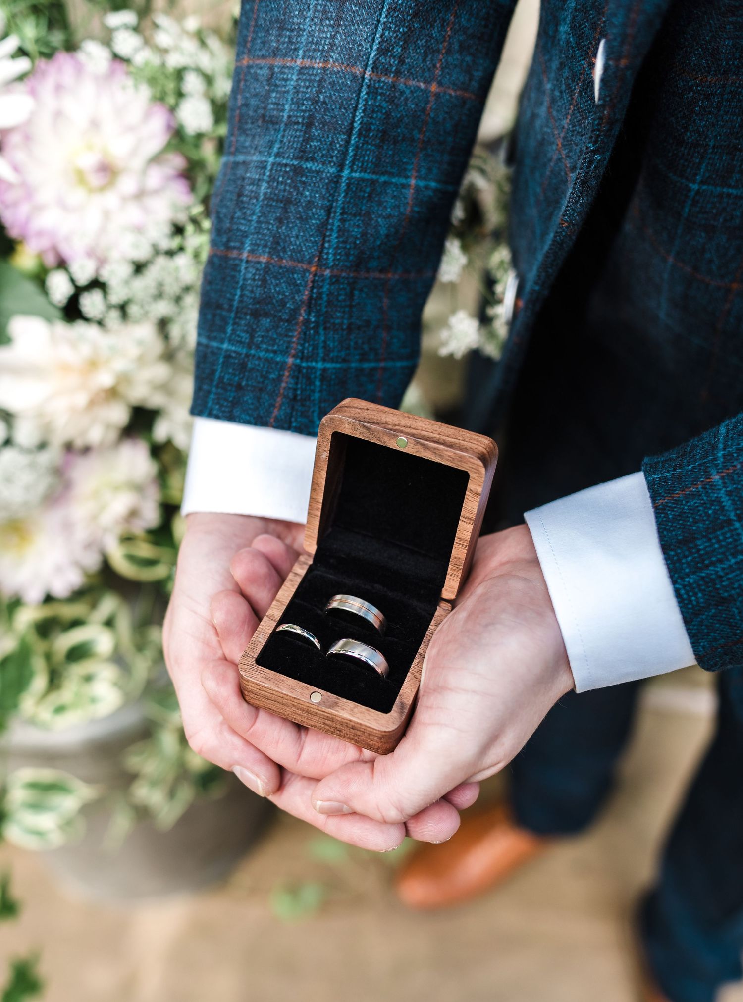 Wedding rings displayed in a wooden box held by hands wearing a navy checkered suit with white cuffs.