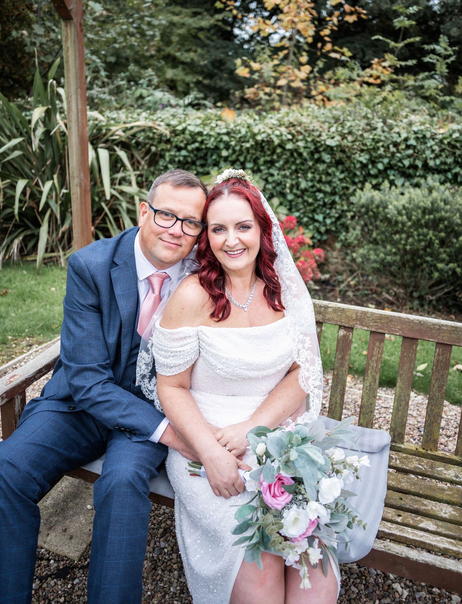A couple sits together on a wooden garden bench with a beautiful floral bouquet.