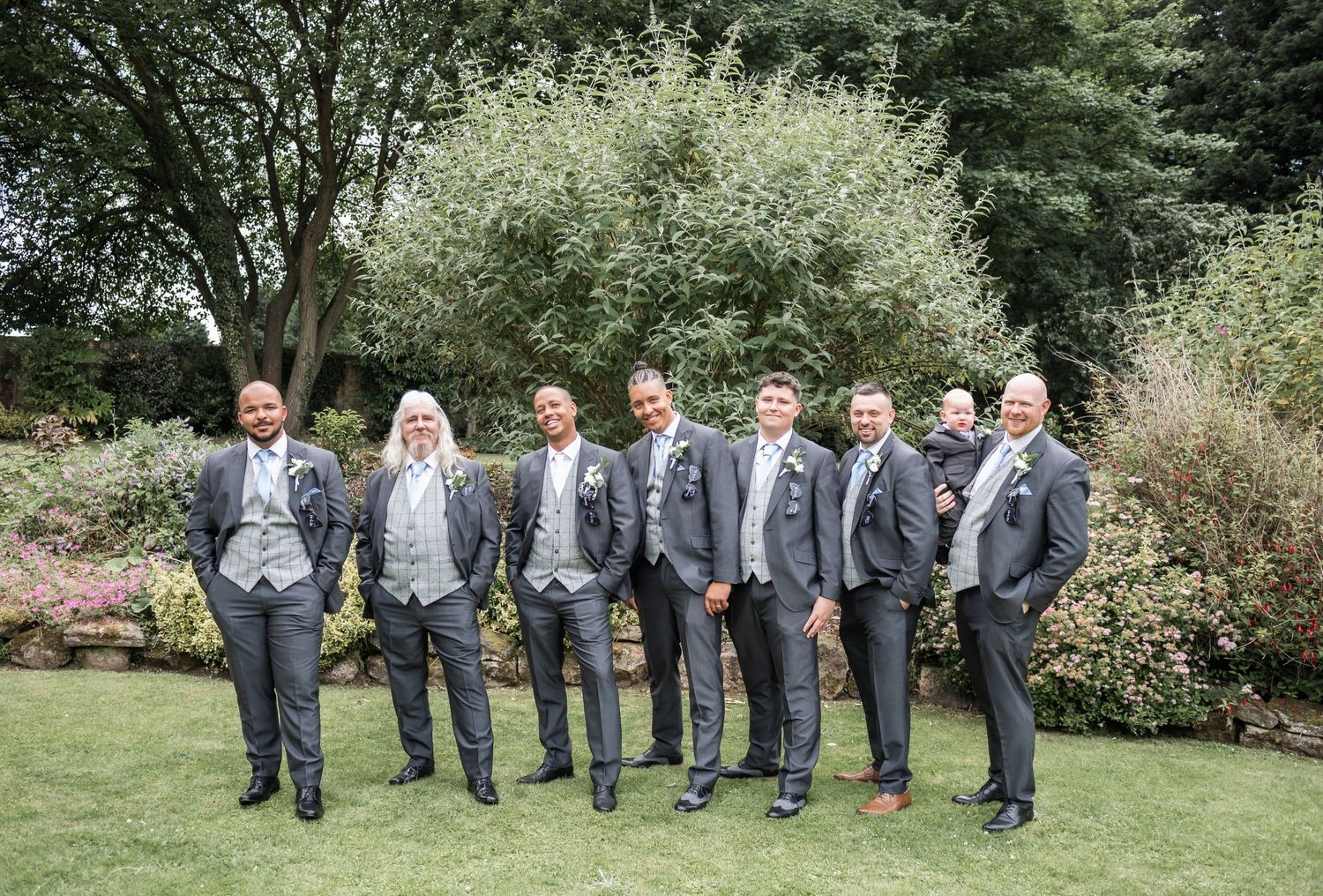 Groomsmen in coordinated grey suits pose together for a formal photo in a lush garden setting.