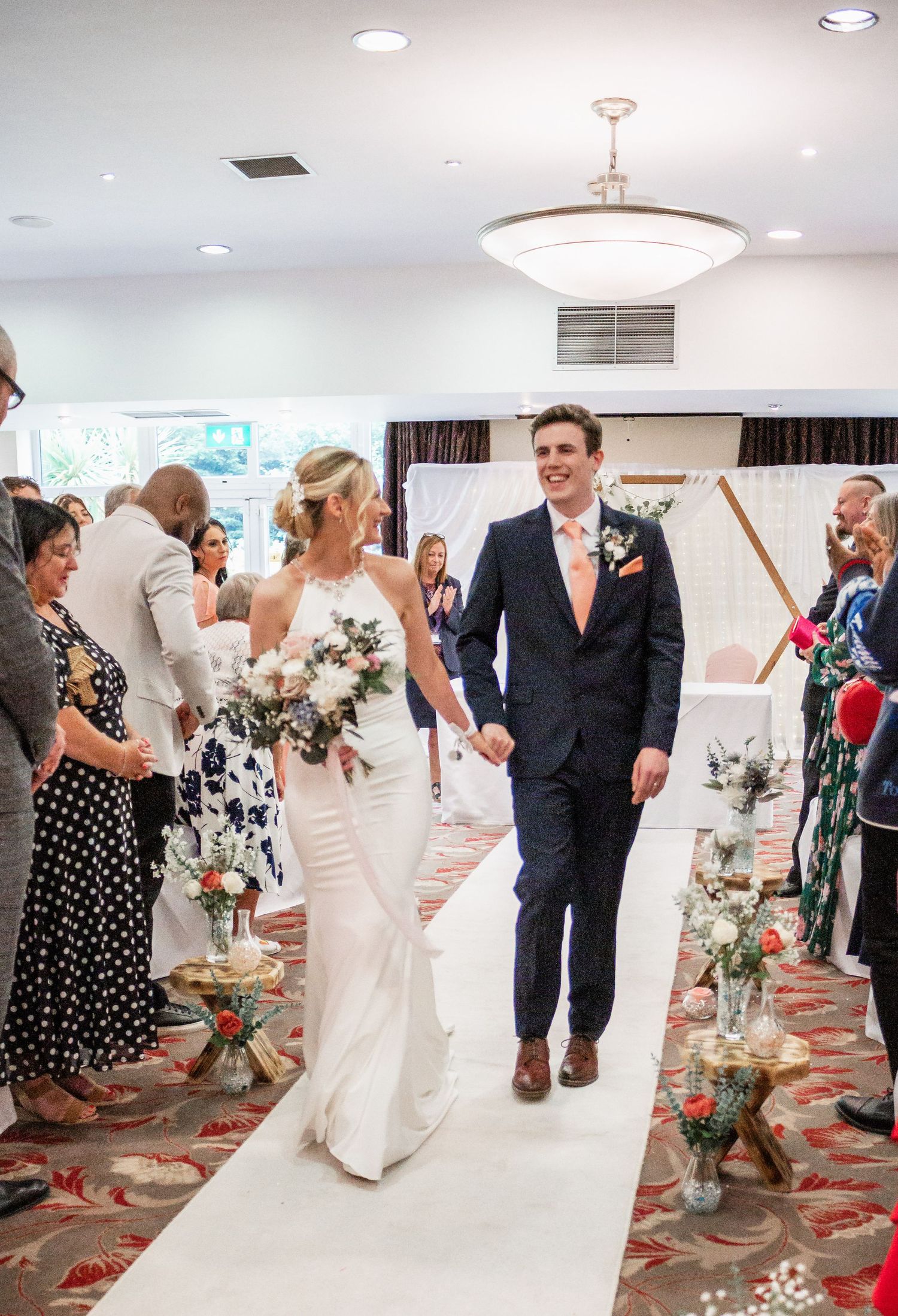 A wedding ceremony in progress with guests lining a white aisle runner decorated with floral arrangements.