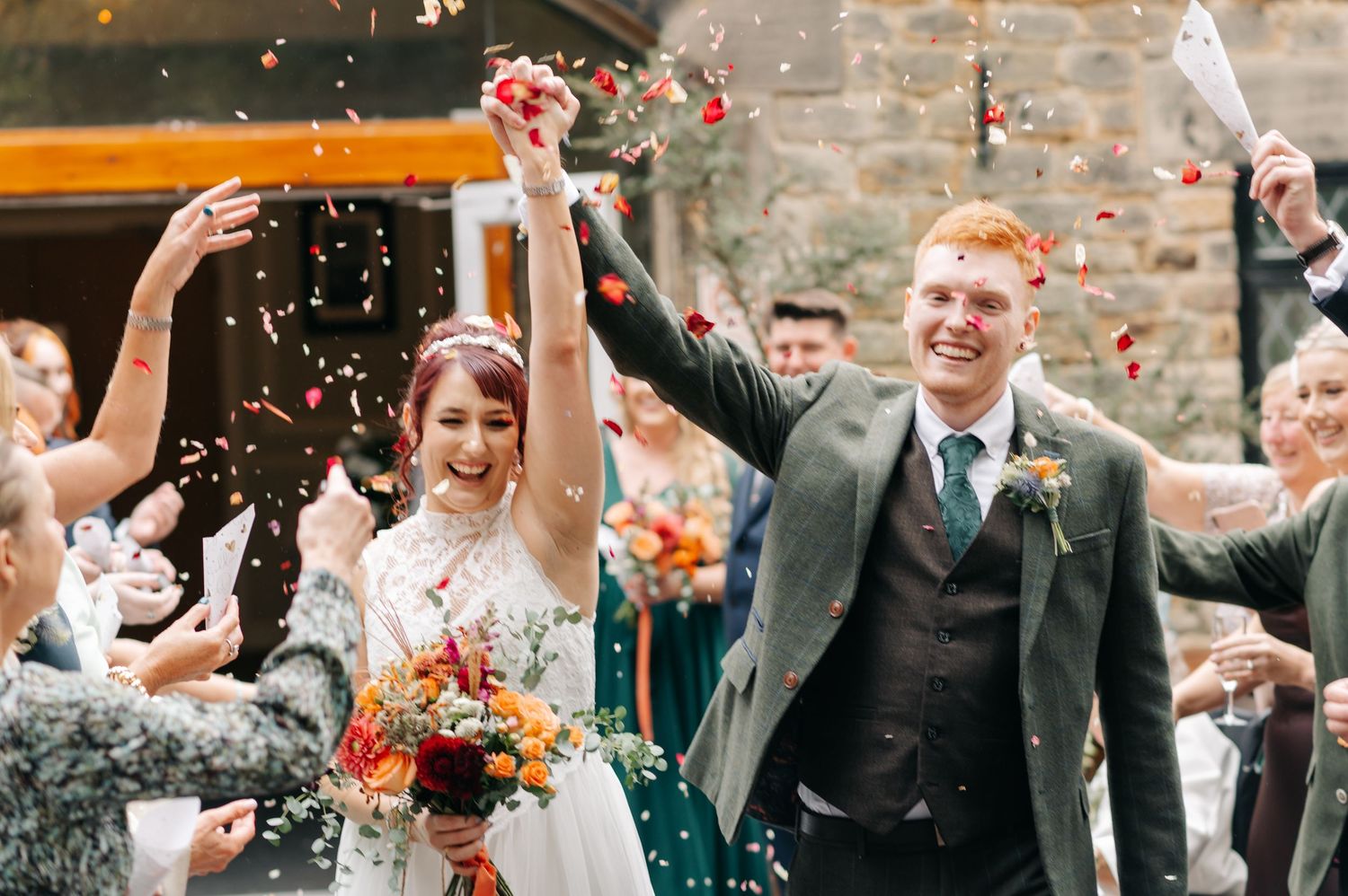 Wedding guests celebrate and throw confetti outside brick venue.