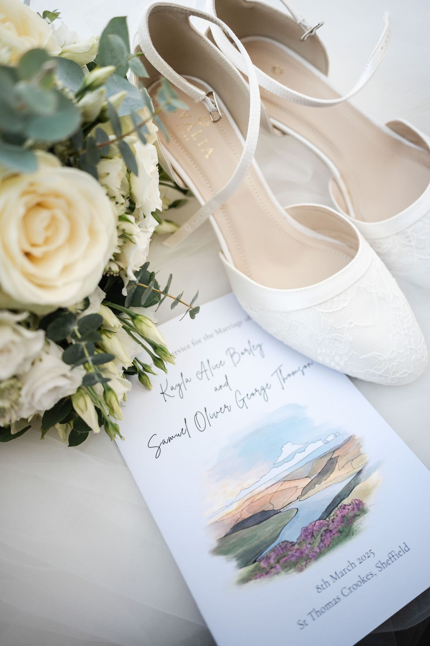 Wedding invitation and white bridal shoes arranged with delicate cream roses and greenery.
