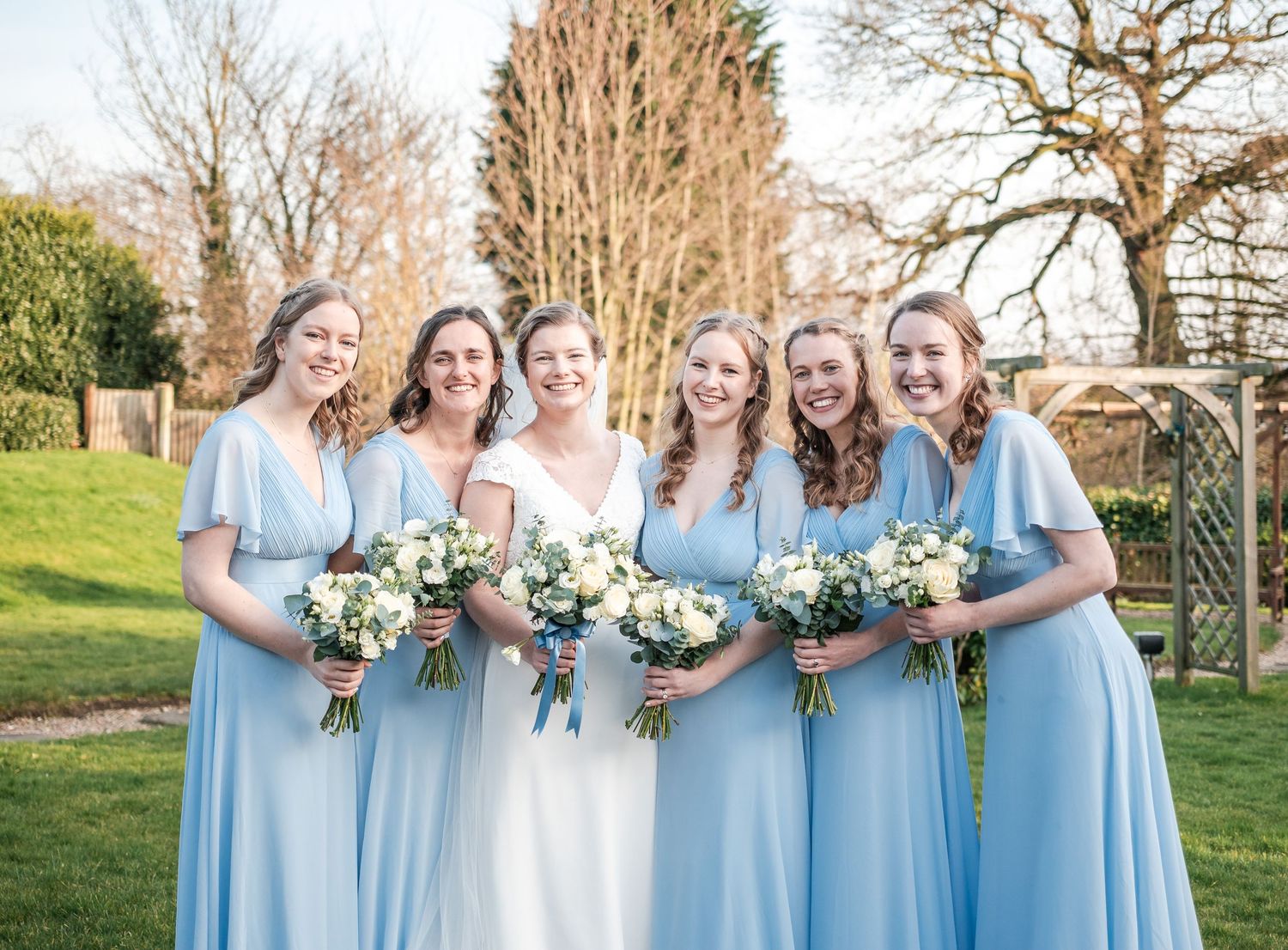 Wedding party in light blue bridesmaid dresses holding white floral bouquets in a garden setting.