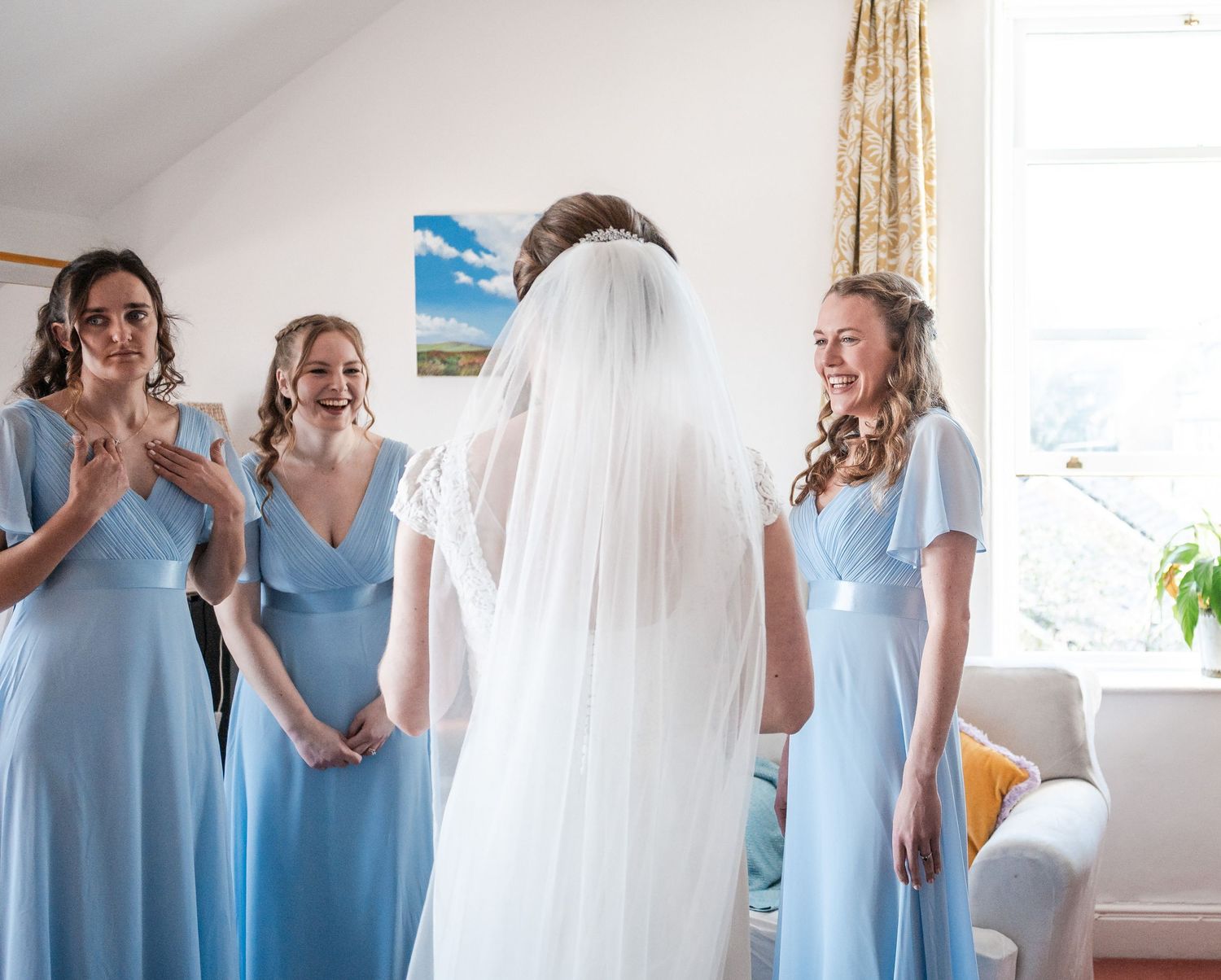 Bridesmaids in light blue dresses smile and react as they see the bride in her flowing white veil.