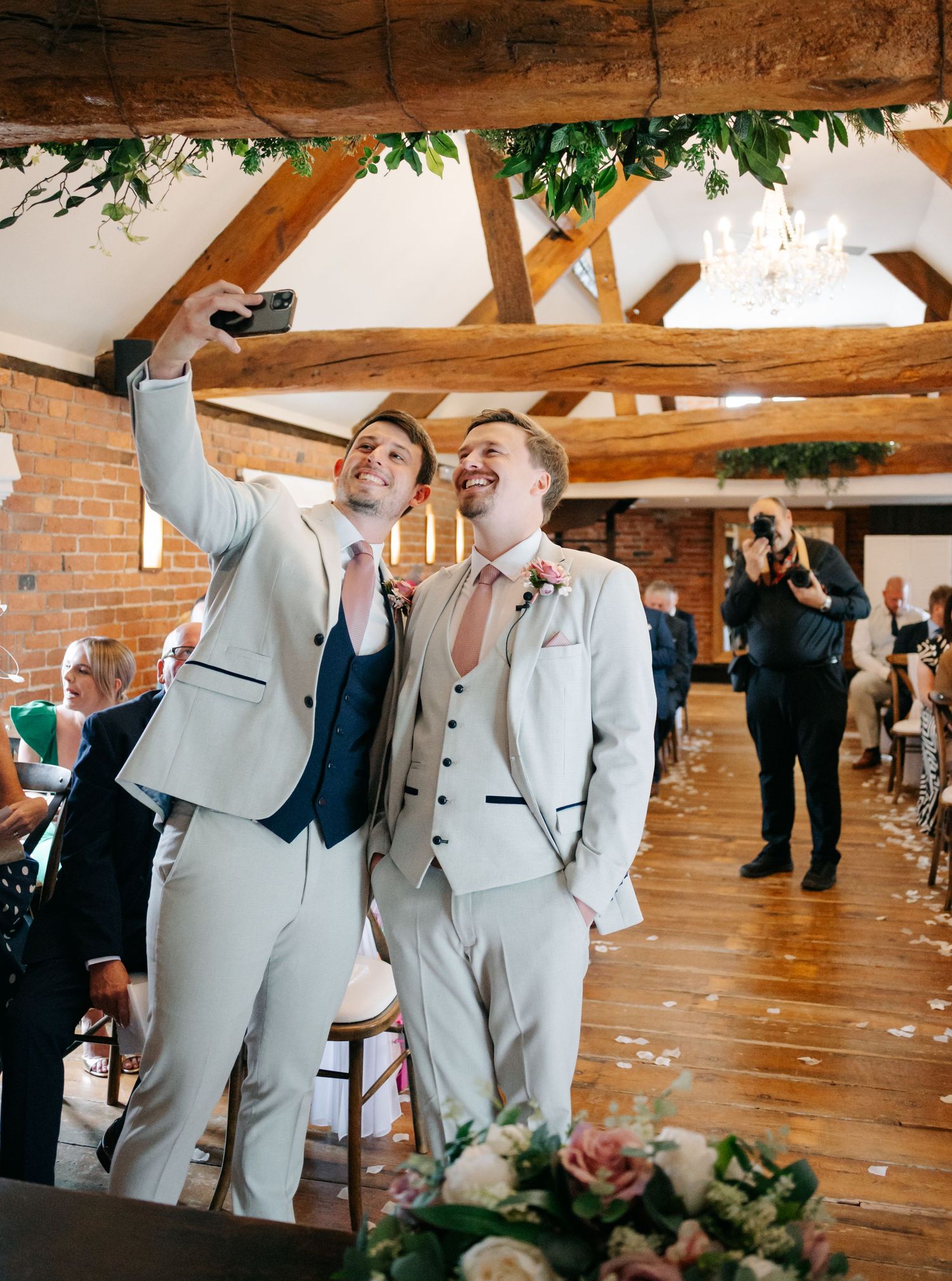 Two people in light grey suits sharing a joyful moment during an indoor wedding ceremony.