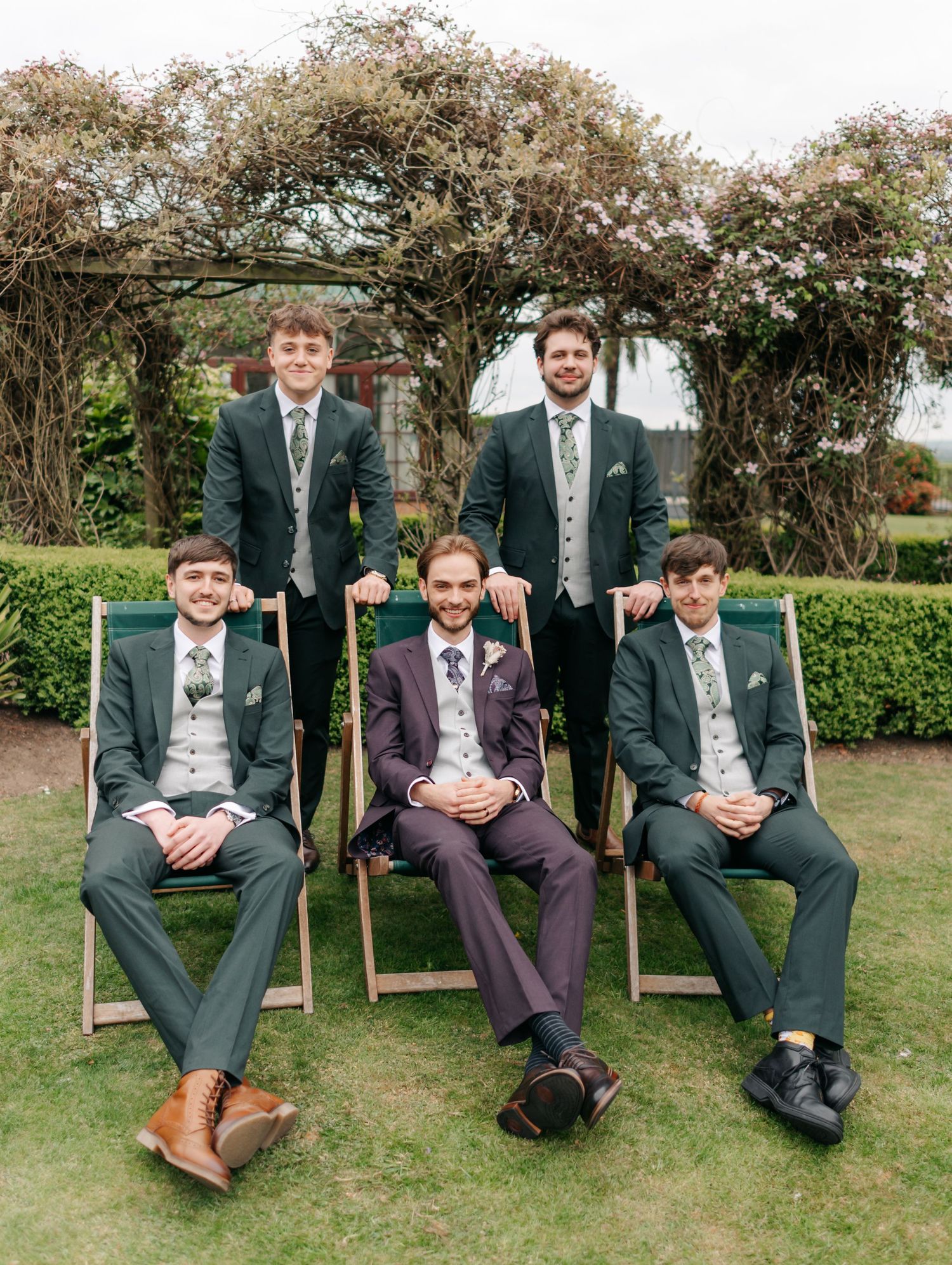 Groomsmen in dark suits relaxing on wooden garden benches outdoors.
