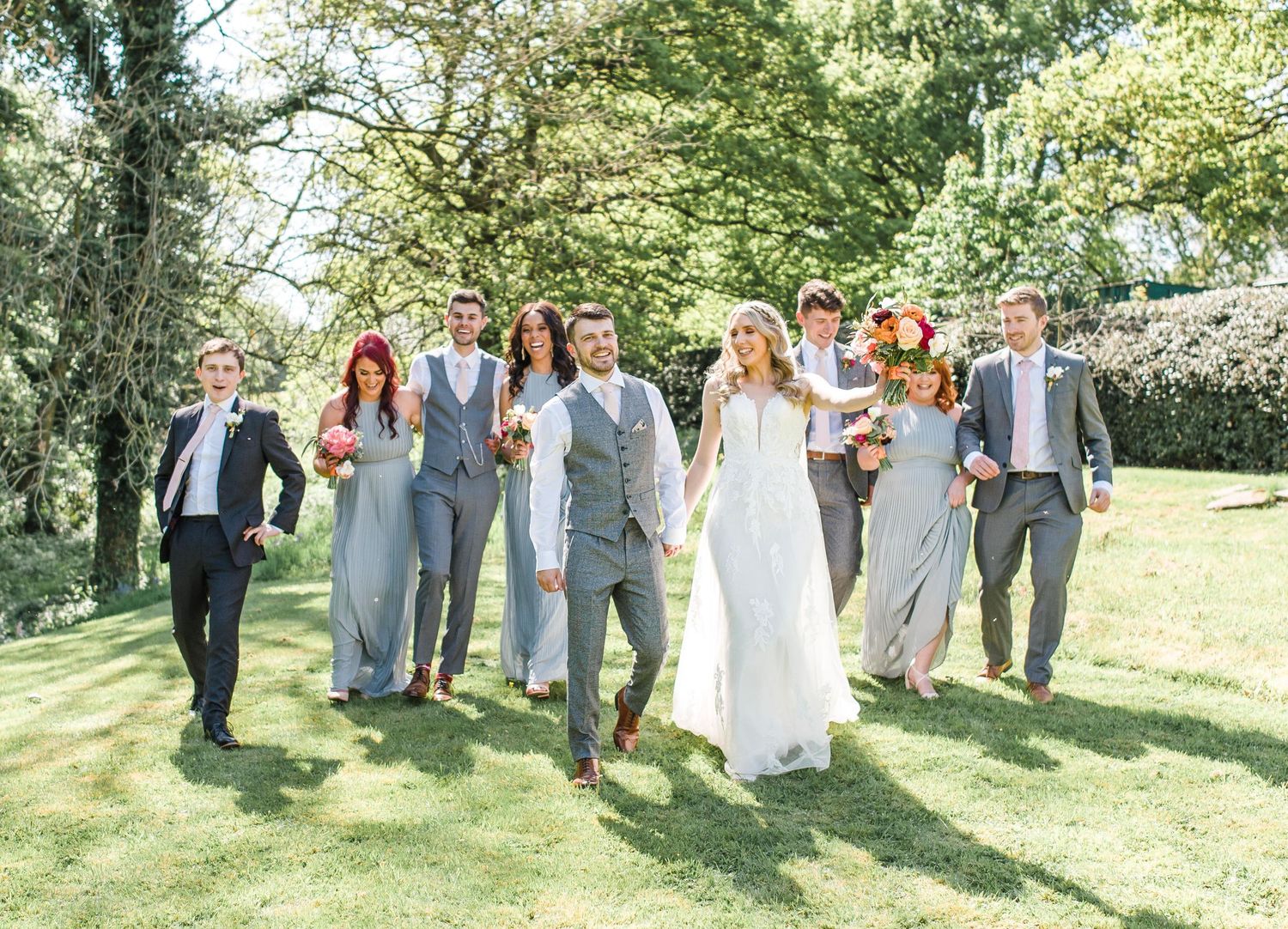 Wedding party walking joyfully together on a sunny lawn with bridesmaids in blue and groomsmen in gray suits.